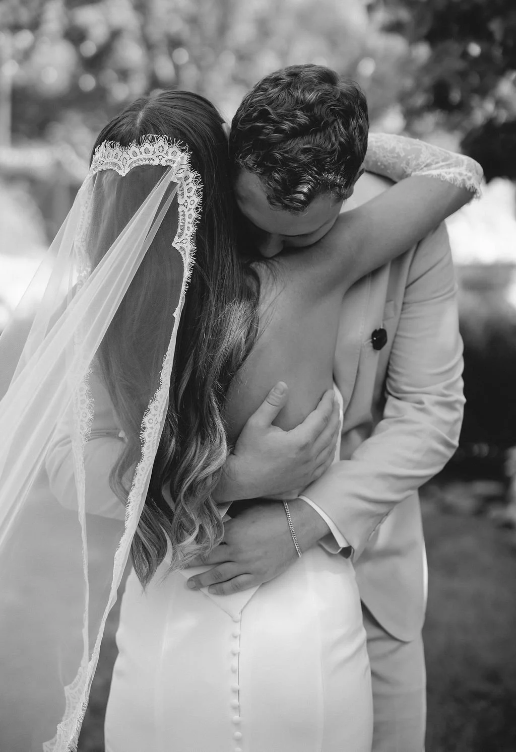 Black and white photo of a couple embracing, with the woman wearing a lace-edged veil and wedding dress, and the man in a light-colored suit.