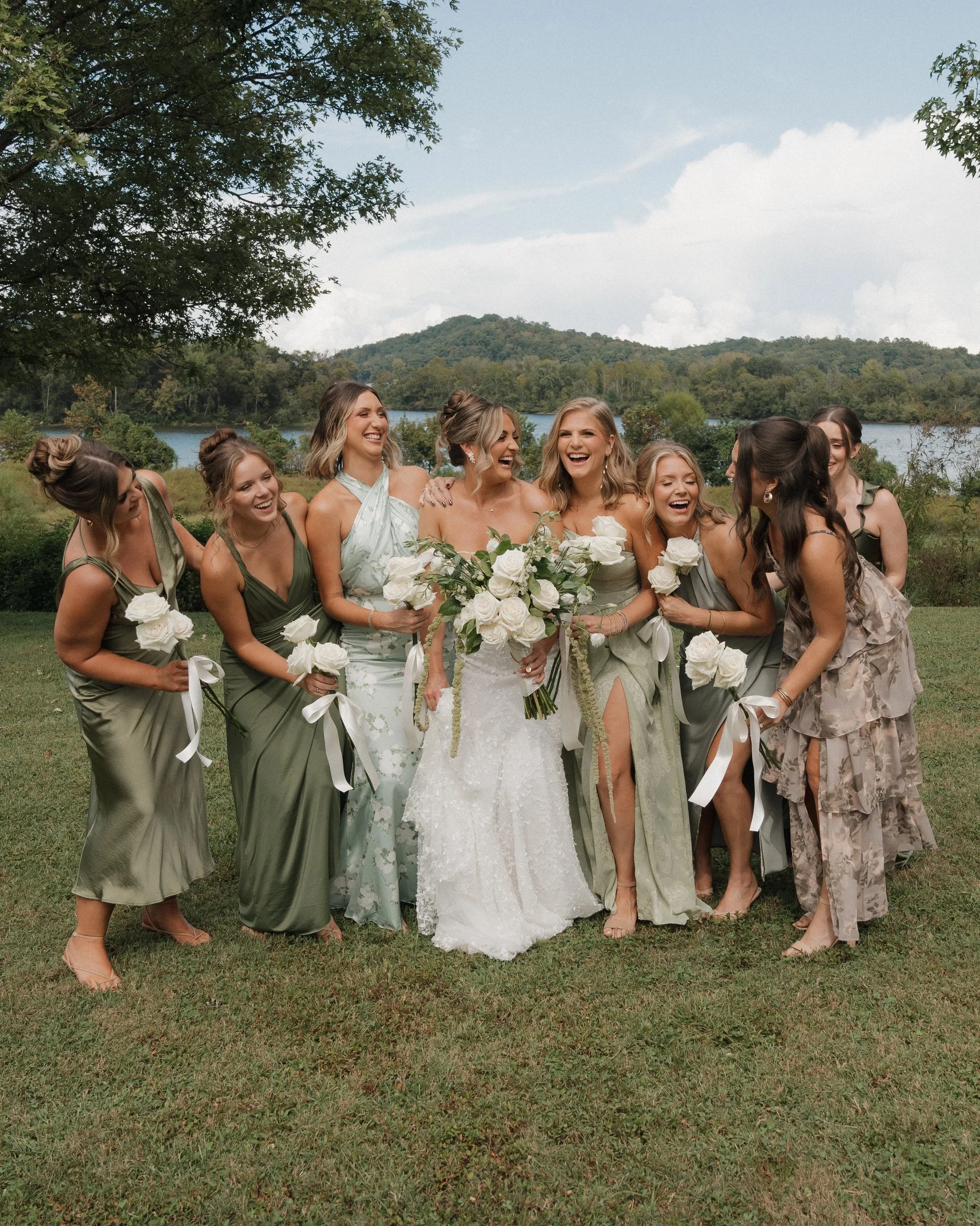A bride and her bridesmaids standing outdoors on grass, holding white roses and laughing. The background features trees, a body of water, and green hills under a partly cloudy sky.