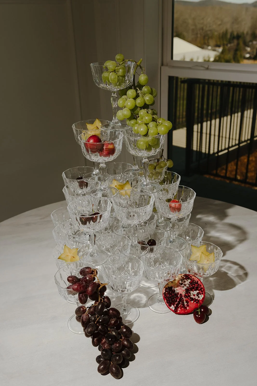 A pyramid of empty crystal glasses filled with grapes, cherries, and starfruit, with black grapes and a halved pomegranate on a white table near a window.