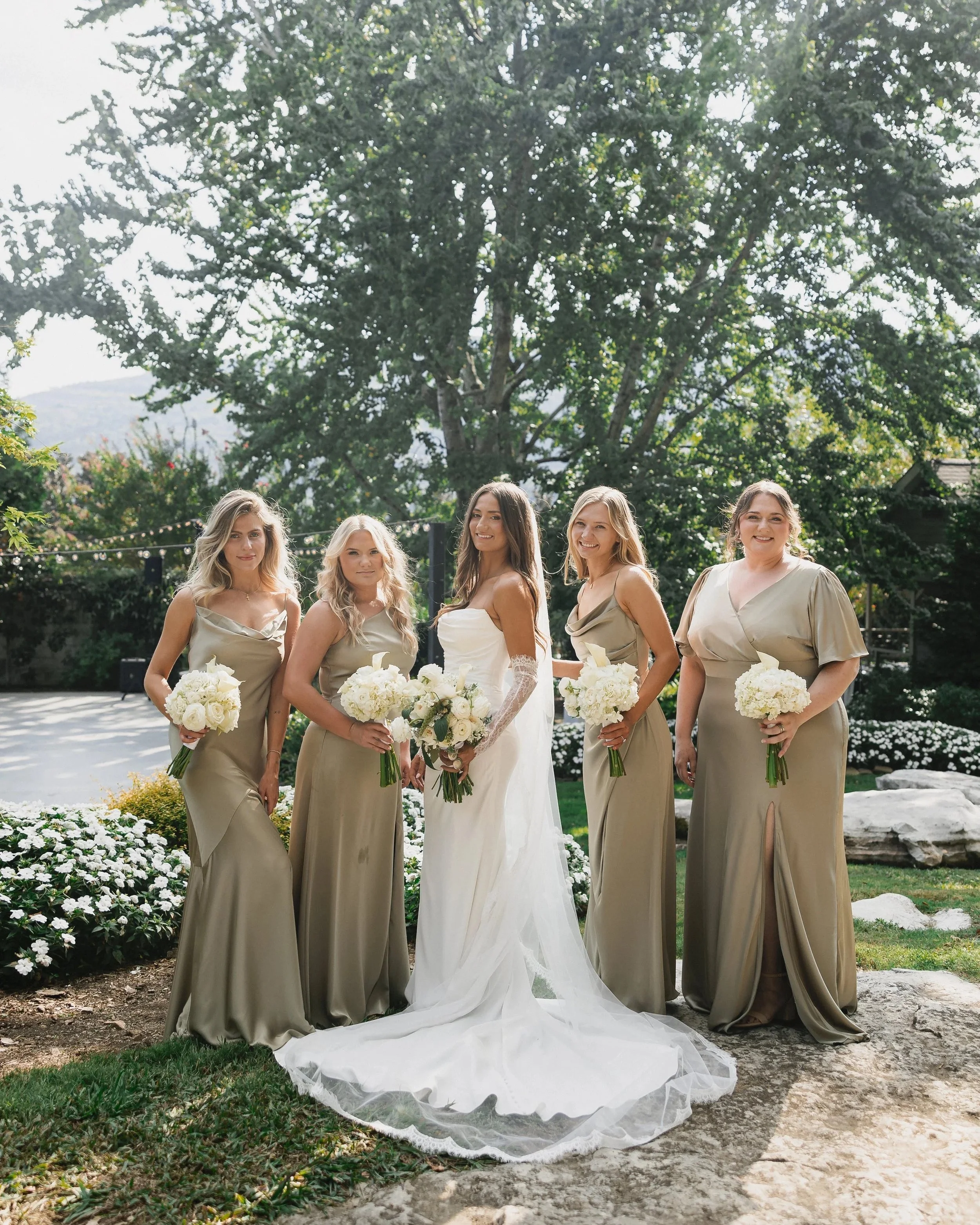 A bride and four bridesmaids standing outdoors with trees and flowers in the background, all holding bouquets of white flowers.