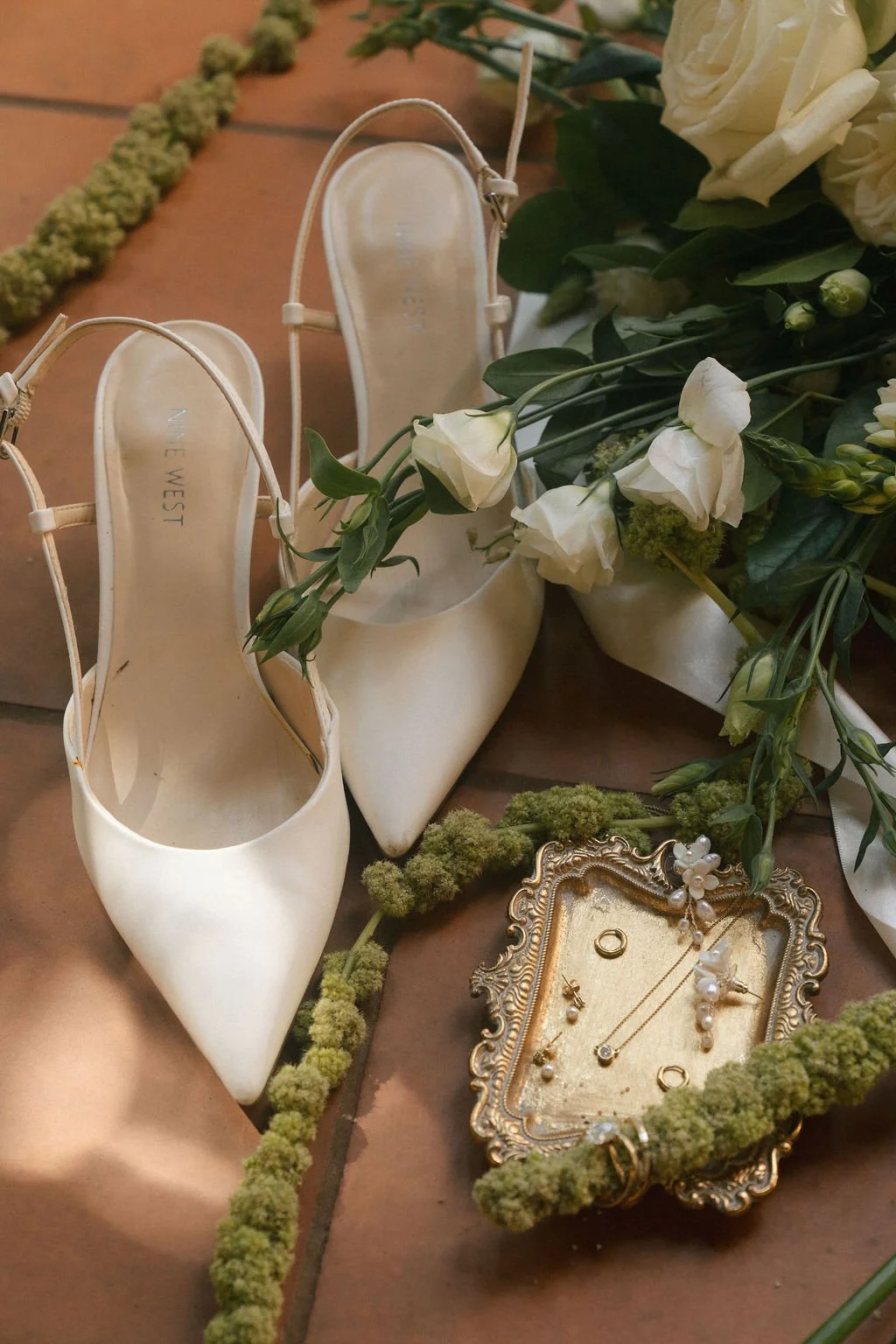 A pair of white high-heeled shoes from Nine West, surrounded by white roses and greenery, with jewelry and wedding rings on an ornate tray, all placed on a tiled floor.