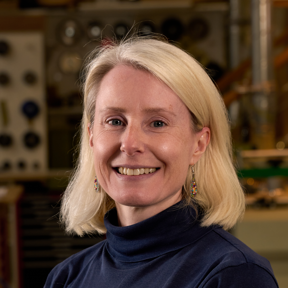 A woman with blonde hair smiling, wearing a navy blue turtleneck and colorful earrings, in a workshop or studio setting.