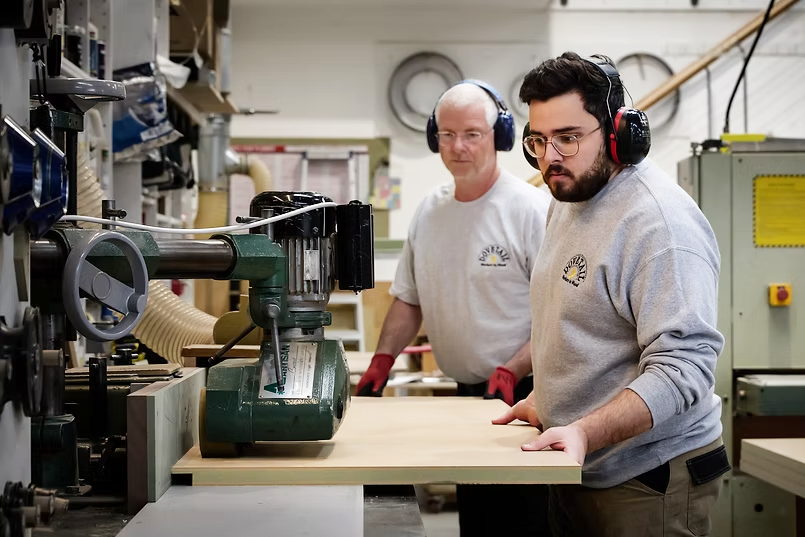 Two men working in a woodworking shop, using a large industrial machine to process a piece of wood. Both are wearing ear protection and casual work clothes.