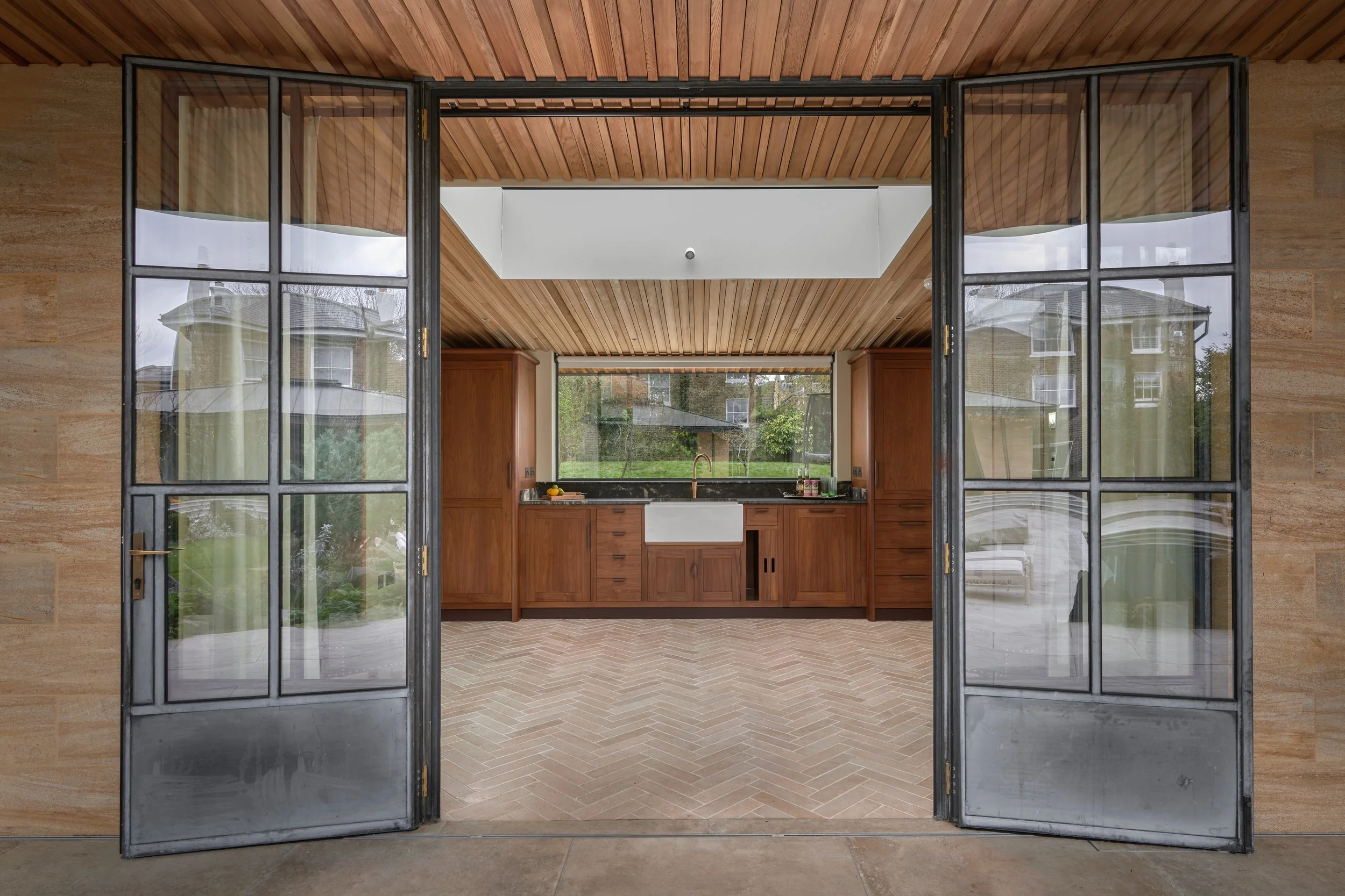 Open black-framed glass doors leading into a kitchen with wooden cabinets, a window above the sink, and a wooden ceiling with a white skylight.