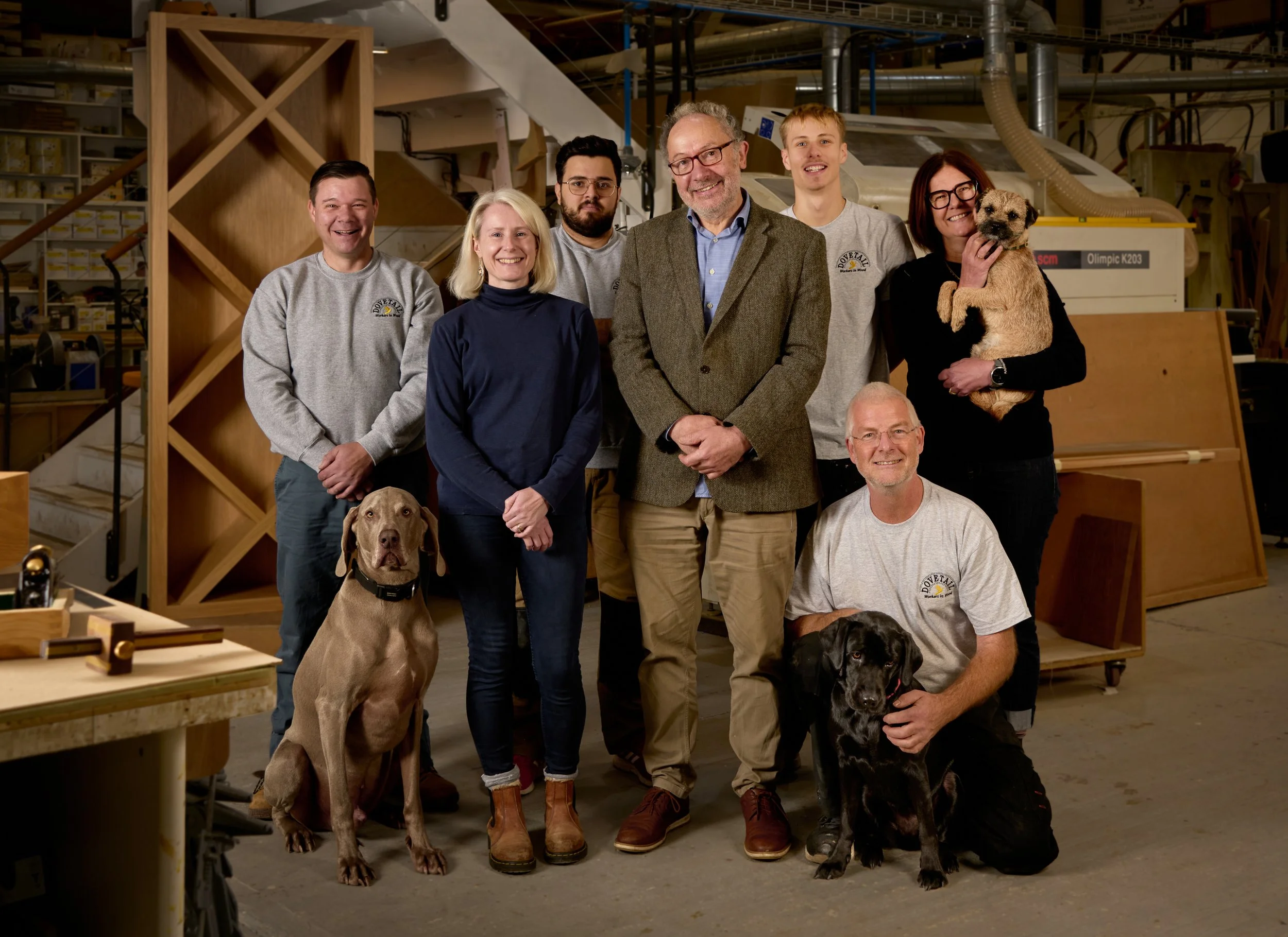 Group of seven people and three dogs standing in a woodworking workshop with shelves and machinery in the background.