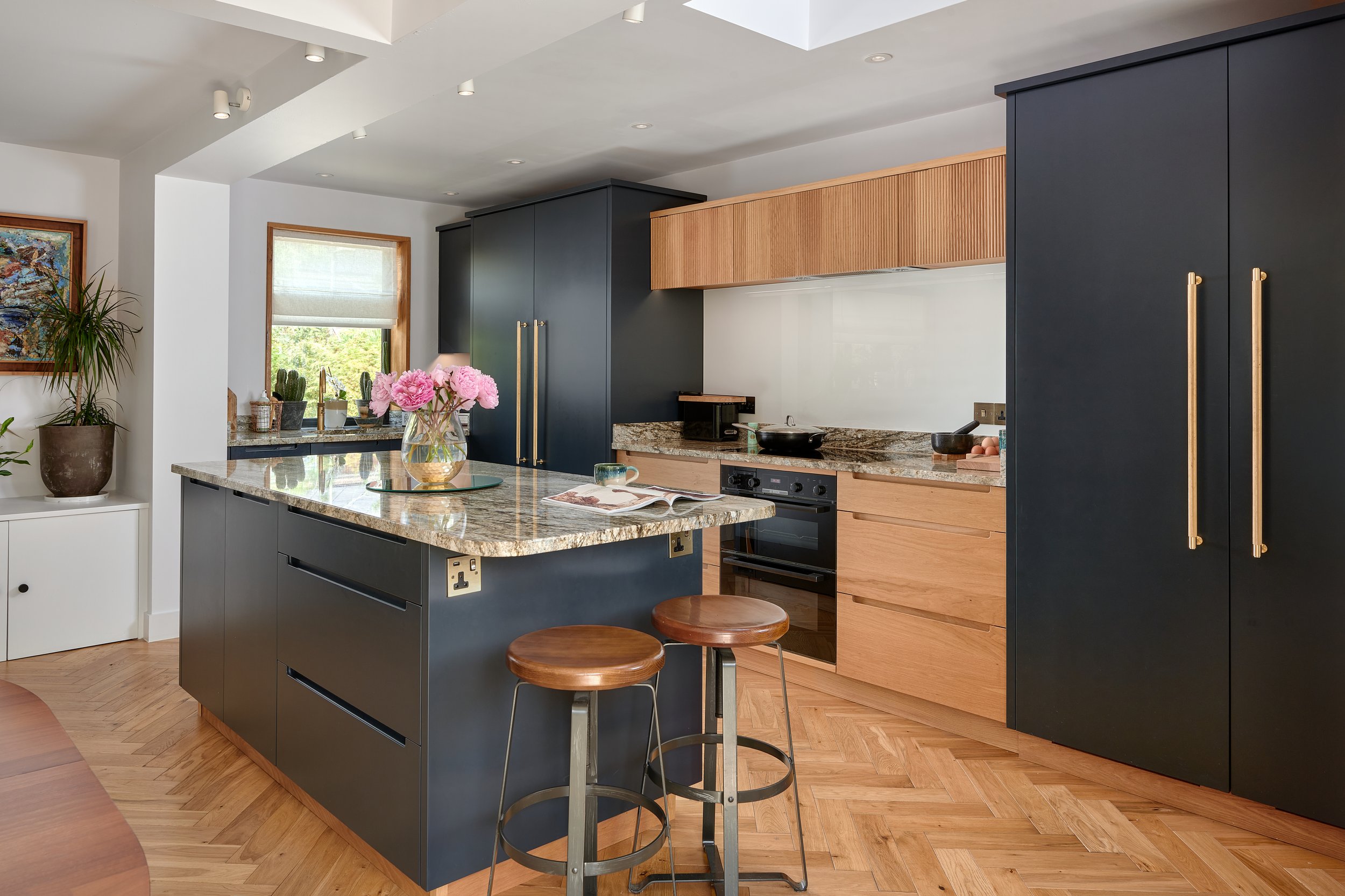 Modern kitchen with navy blue cabinets, wooden drawers, a granite countertop, a kitchen island with a vase of pink flowers, and bar stools with wooden seats. There are trees visible through the window.