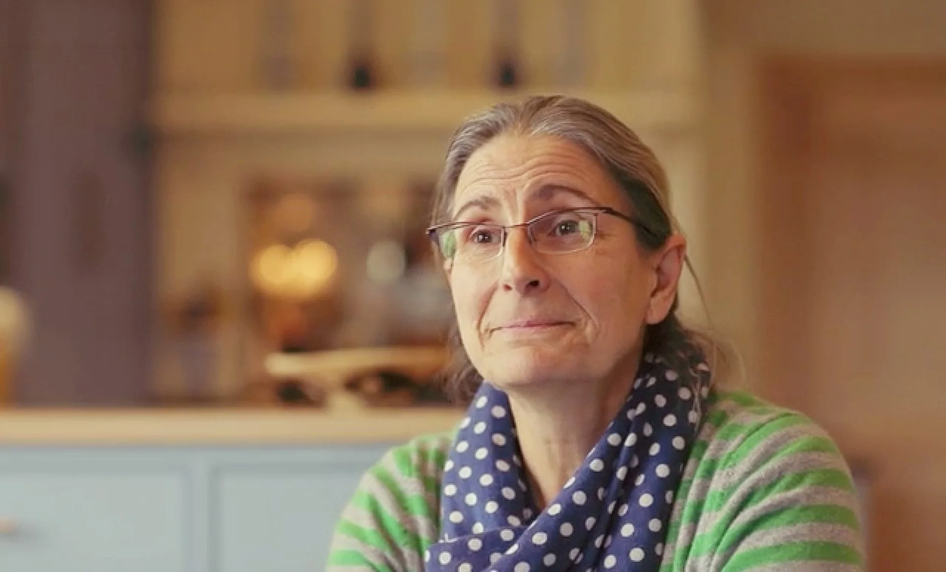 An elderly woman with glasses, wearing a blue polka-dotted scarf and a green striped sweater, smiling softly in a warmly lit indoor setting.