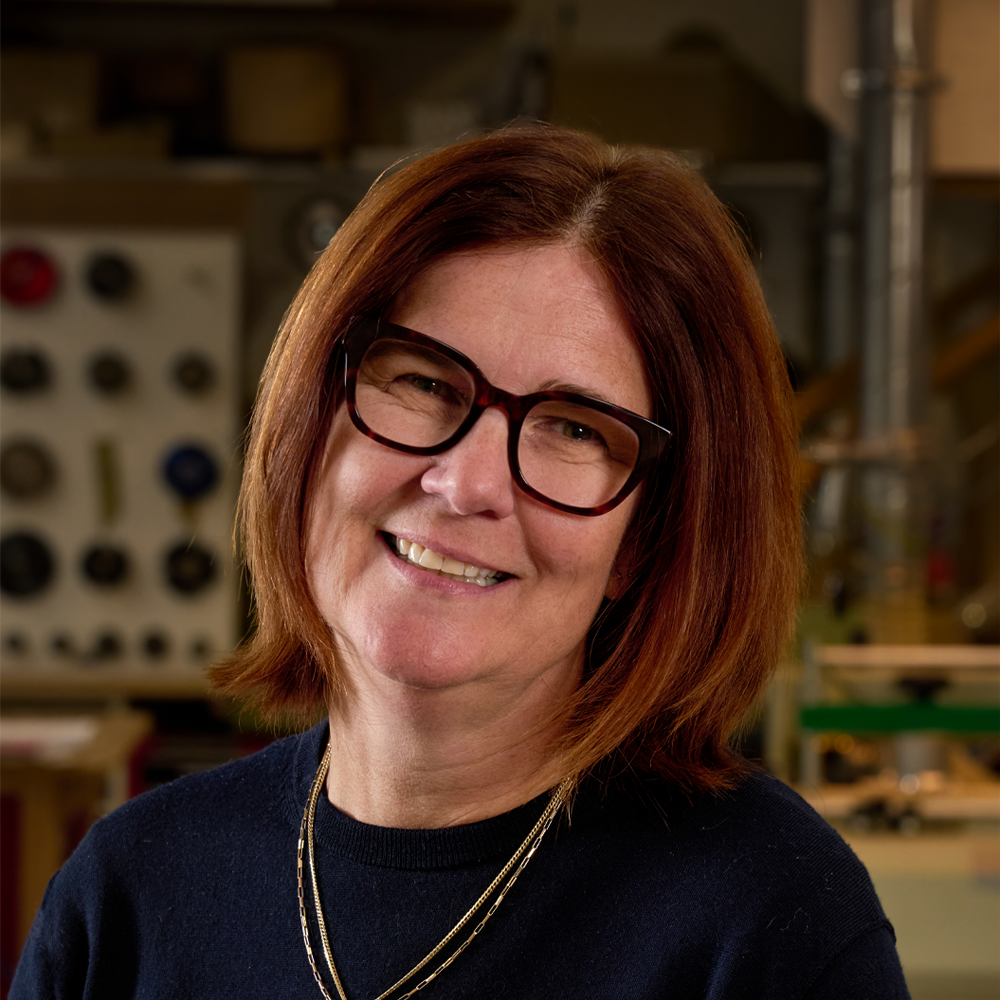 A smiling woman with reddish-brown hair, wearing glasses and a black sweater, in a workshop setting with tools and equipment in the background.