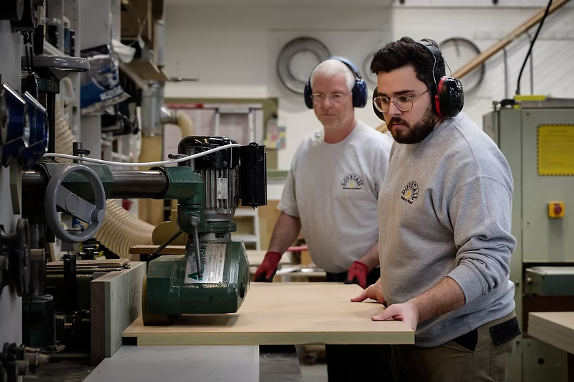 Two men in a woodworking shop, wearing hearing protection, working on a large piece of wood with a woodworking machine.