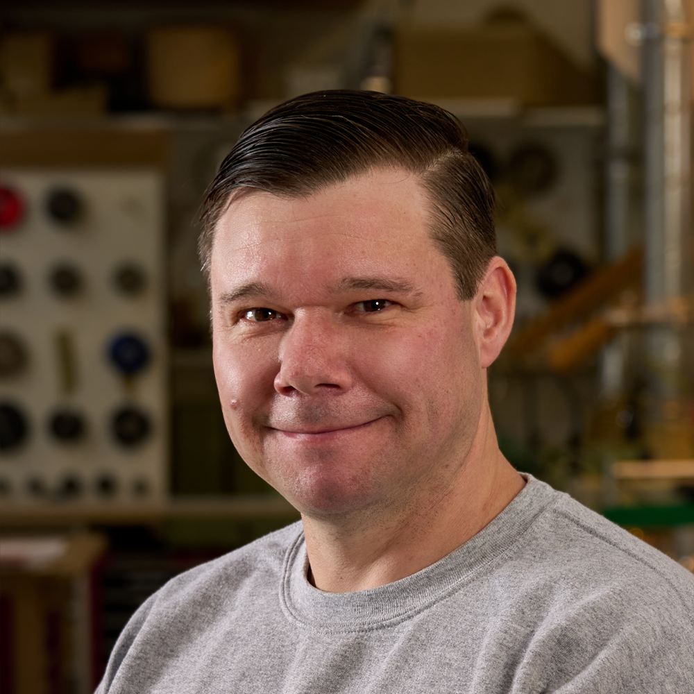 A man with short, slicked-back dark hair wearing a gray t-shirt, smiling in a workshop or garage setting with tools and equipment in the background.