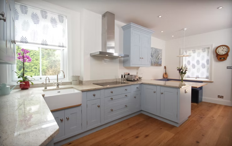 Bright kitchen with light blue cabinets, white granite countertops, a large window with patterned blinds, a built-in sink, and hardwood floors. Decor includes a pink orchid, a small vase with flowers, a wall clock, and a guitar.