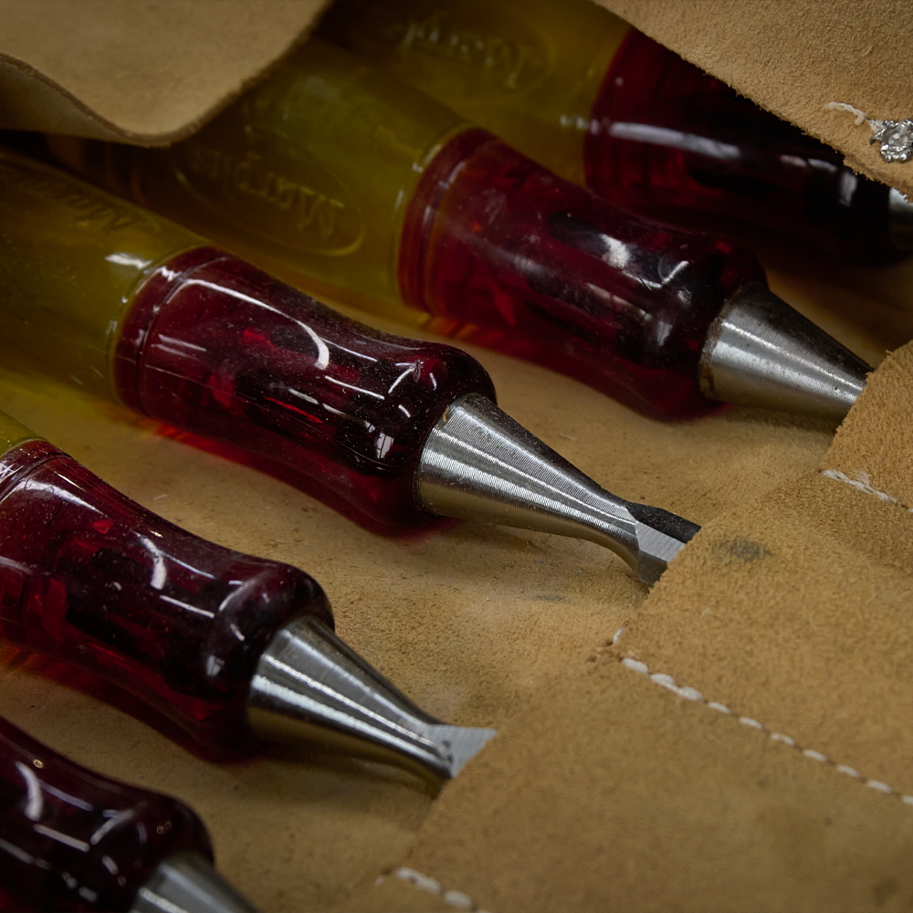 Close-up of several sewing needles with red handles resting on a tan leather fabric with white stitching.