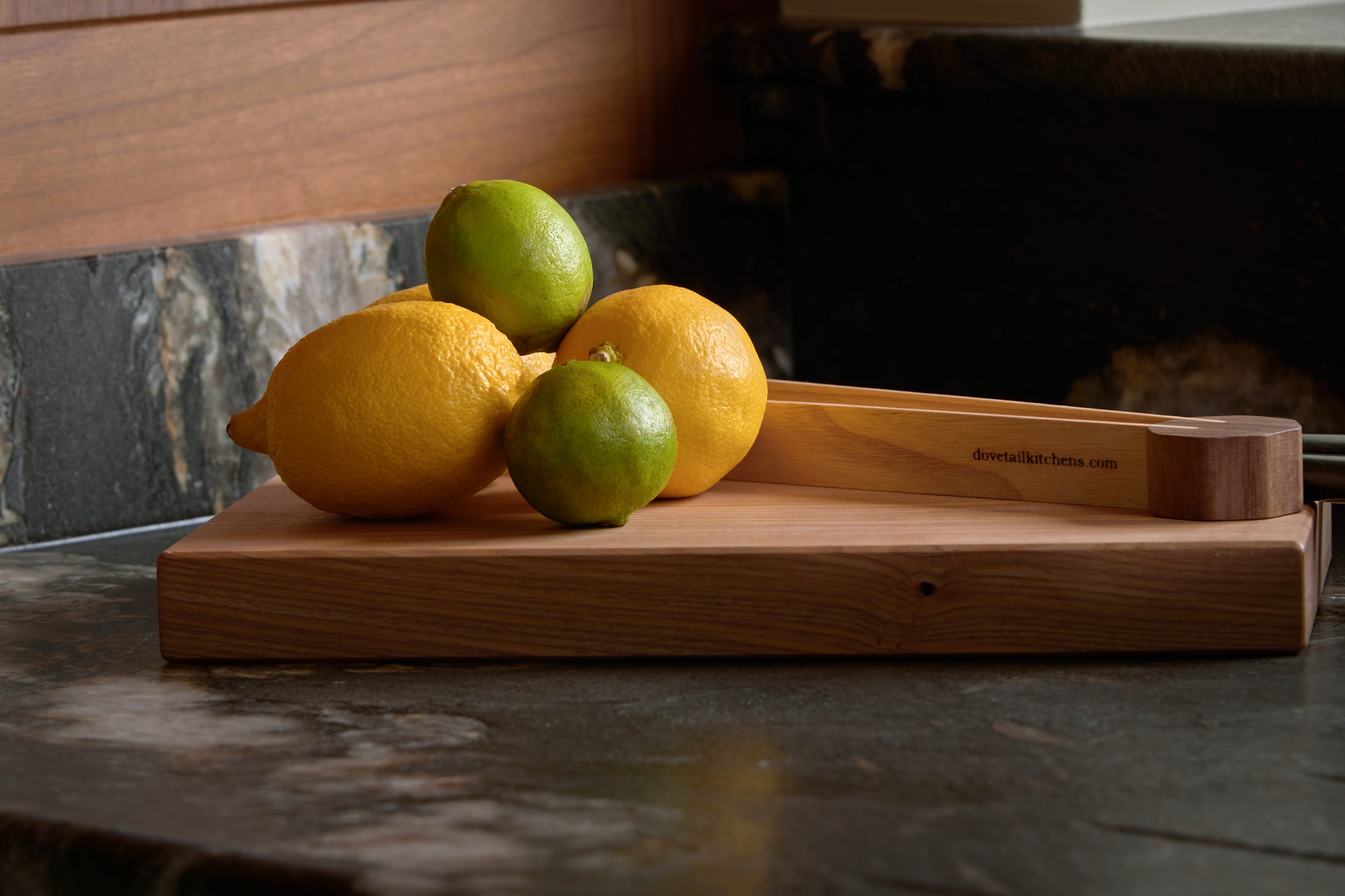 A wooden cutting board with two lemons and two limes on a dark stone countertop in a kitchen.