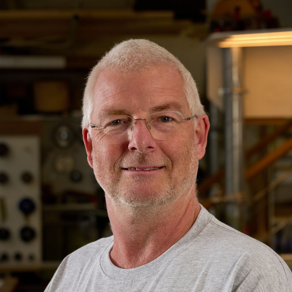 Portrait of an older man with glasses, short gray hair, and a trimmed beard, smiling, in a workshop.