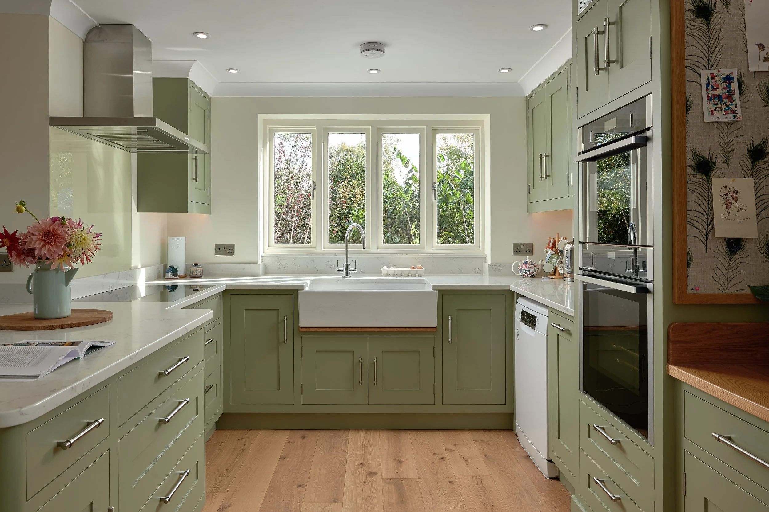 Bright kitchen with green cabinets, a large window above the sink showing greenery outside, and various kitchen appliances and decorations.