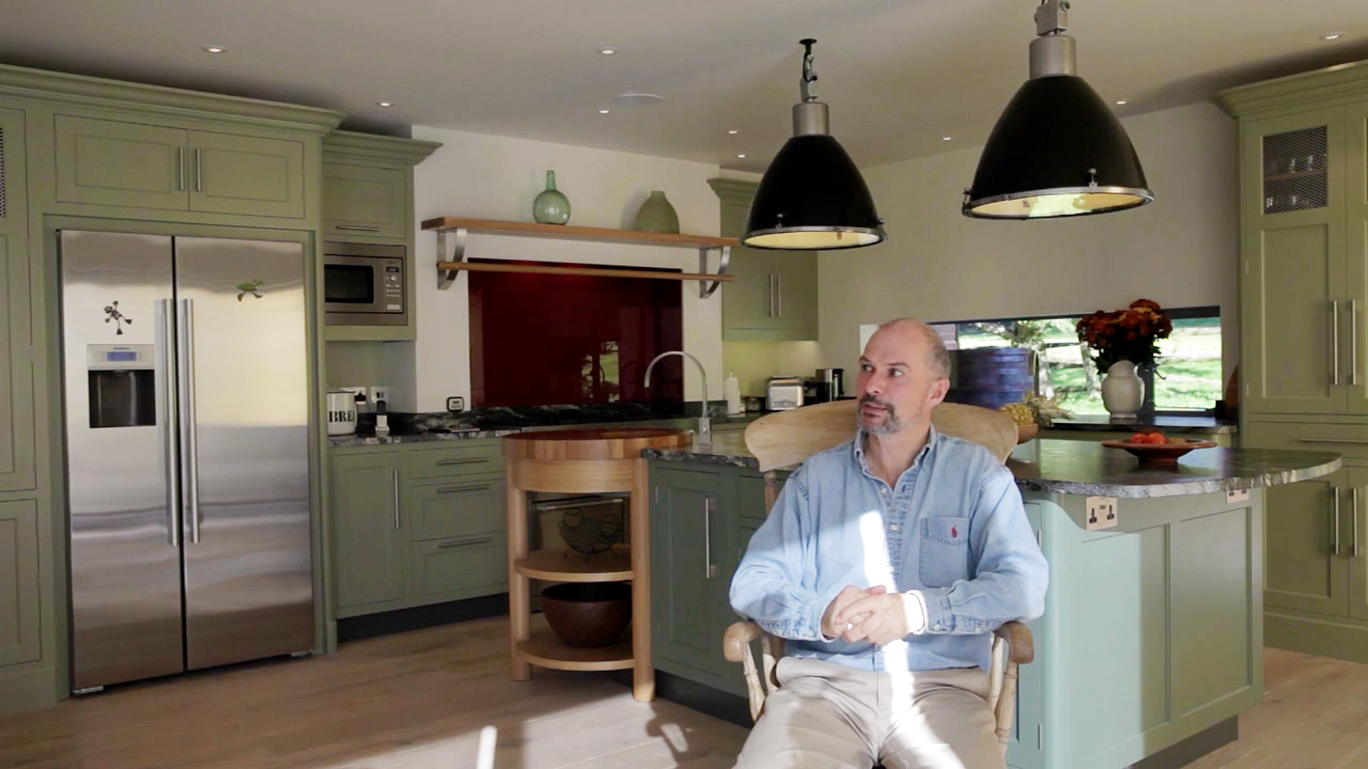 A man with a beard and light blue shirt sitting in a wooden chair in a kitchen with green cabinets, a stainless steel refrigerator, and pendant lights. There is a window with a vase of flowers, a bowl on the counter, and kitchen appliances in the background.