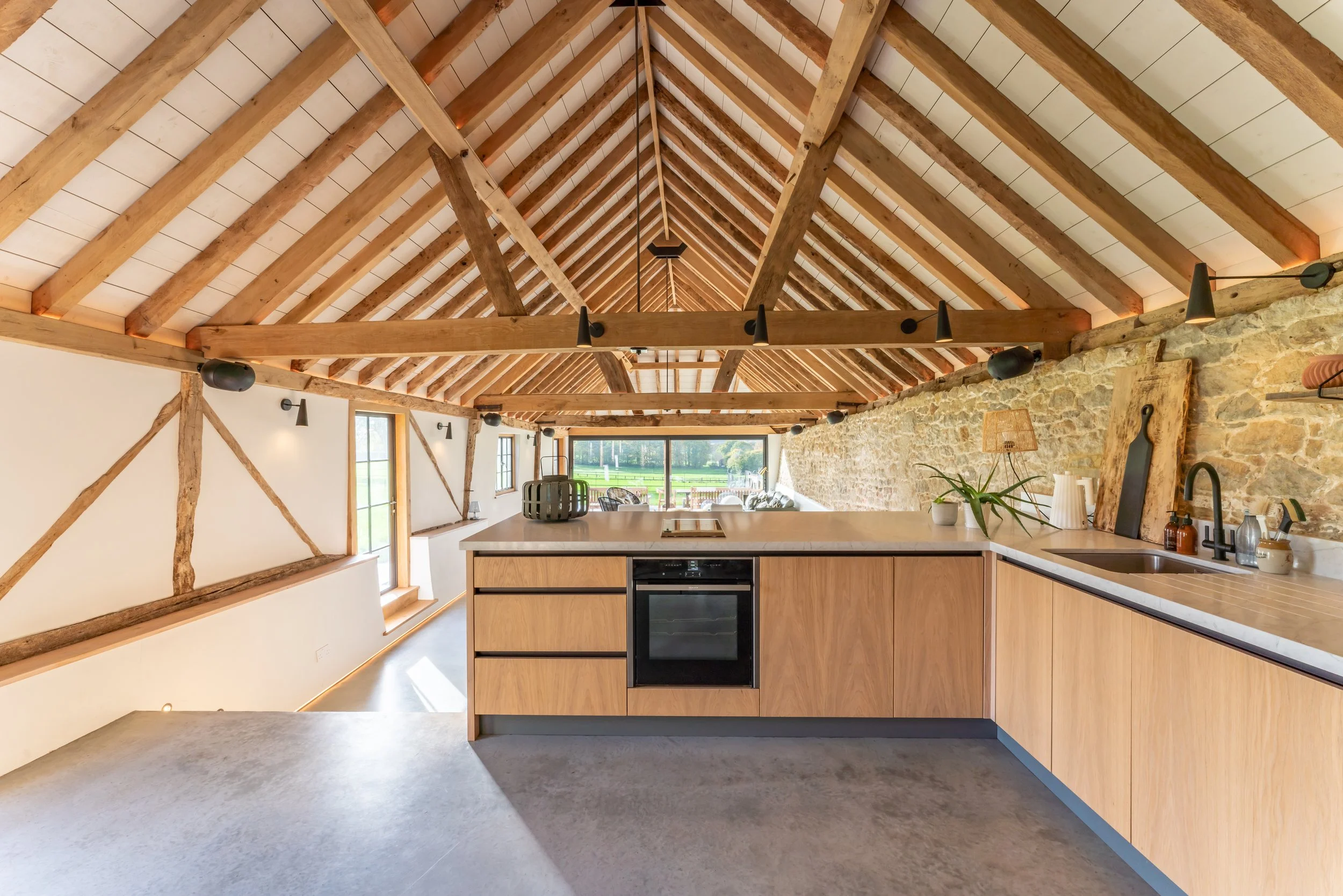 Open-concept kitchen with a vaulted wooden ceiling, exposed wooden beams, a large window with a view of green fields, and modern kitchen cabinets with a built-in oven.