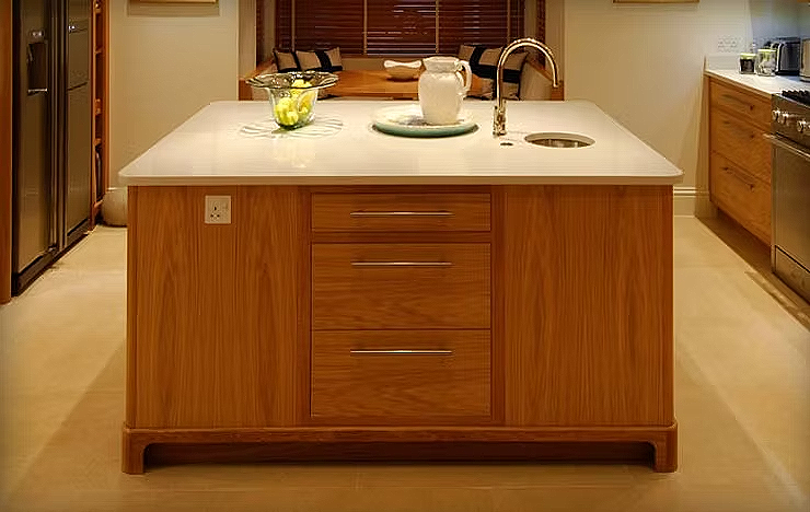 Modern kitchen island with wooden cabinet, white countertop, sink, and decorative items, with a dining area in the background.