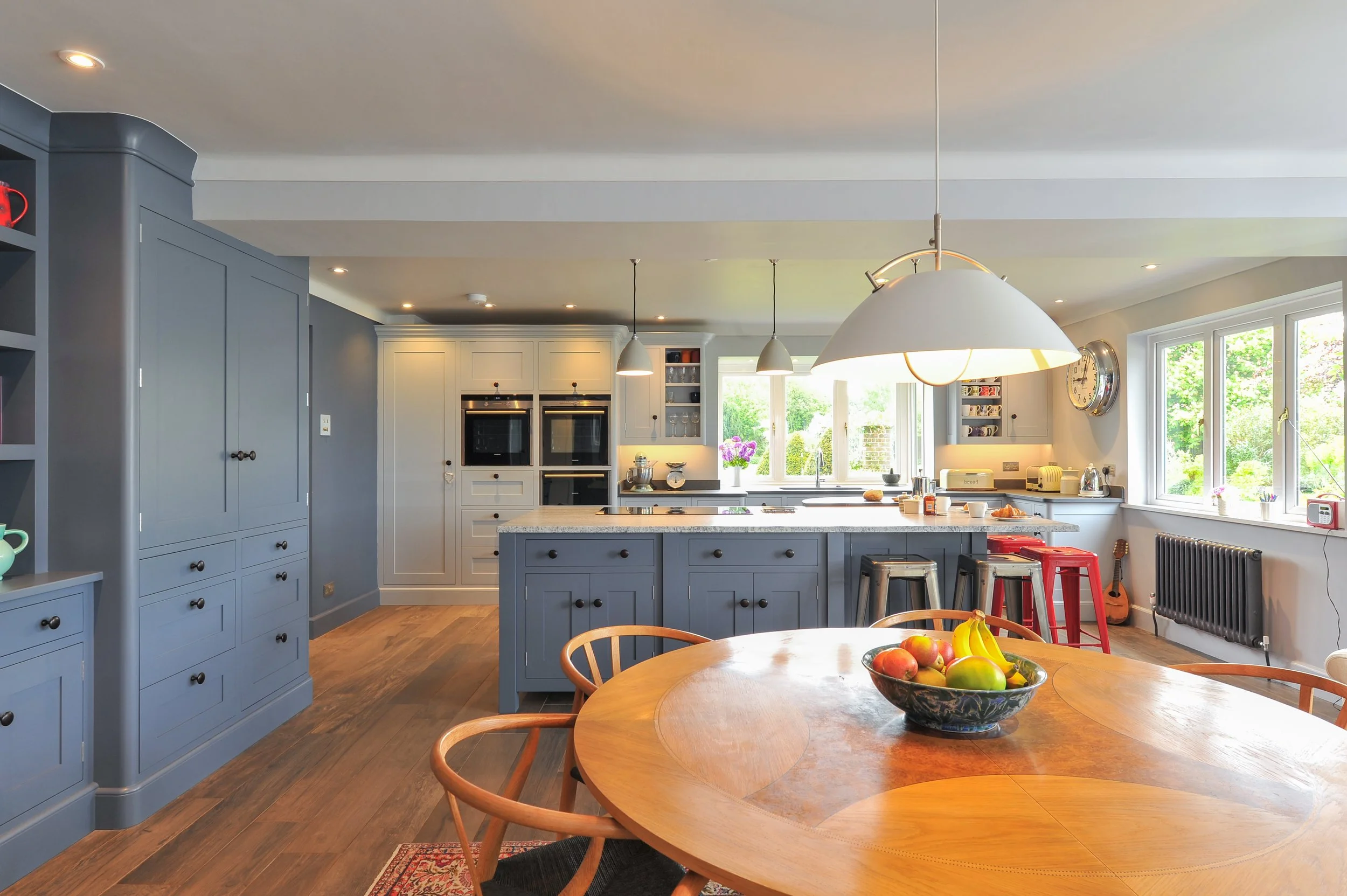Bright kitchen with white cabinets, gray island, wooden dining table with fruit bowl, and large windows showing greenery outside.