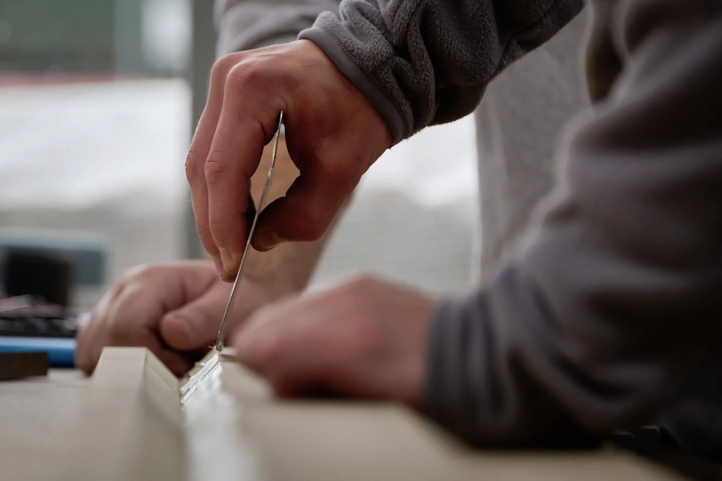Close-up of a person making a model airplane, holding an airplane wing with their left hand and using a fine tool with their right hand. The person is wearing a gray sweater.