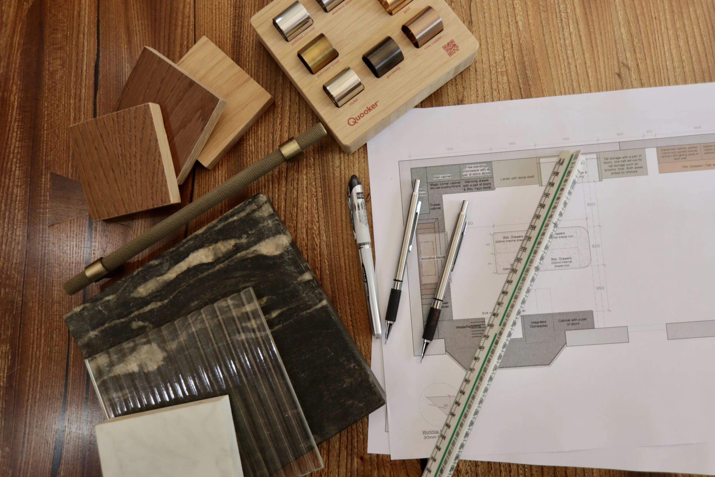 Desk with wooden furniture samples, architectural blueprints, pens, and measuring tools.