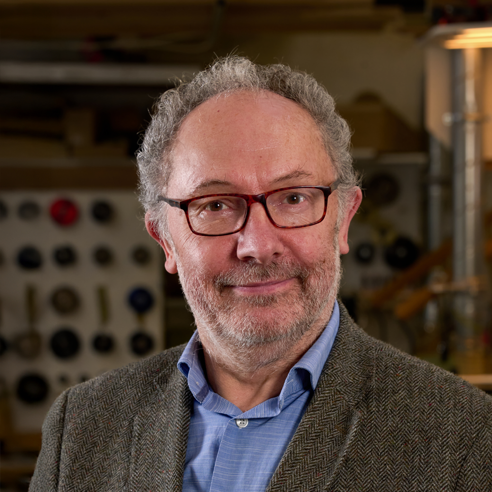 Portrait of a middle-aged man with glasses, gray hair, a beard, wearing a blazer and blue shirt, standing in a workshop with tools in the background.
