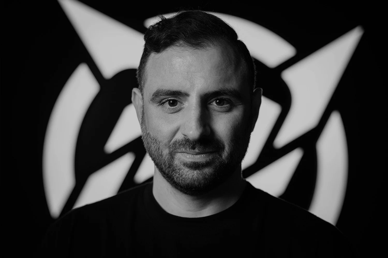 A black and white portrait of a man with short hair and a beard, facing forward with a slight smile, standing in front of an abstract background.