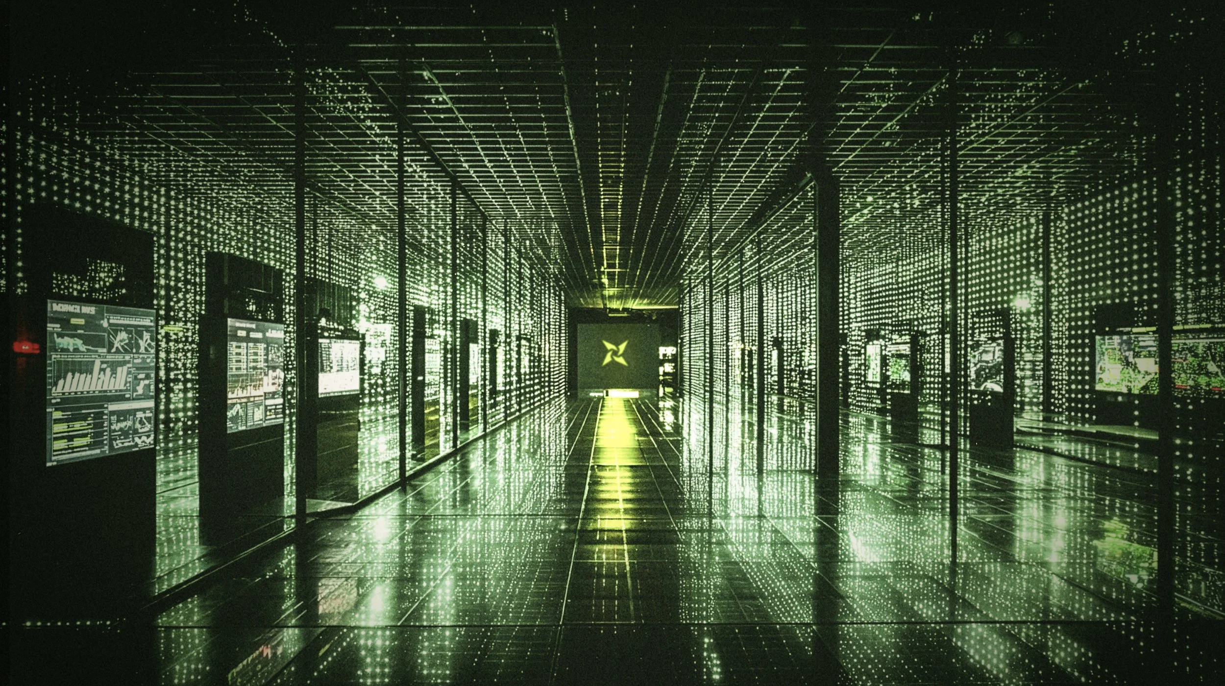 A futuristic digital display room with walls, ceiling, and floor covered in green LED-like lights creating a grid pattern, with multiple screens on both sides showing various charts and data, and a central illuminated logo at the end of the corridor.