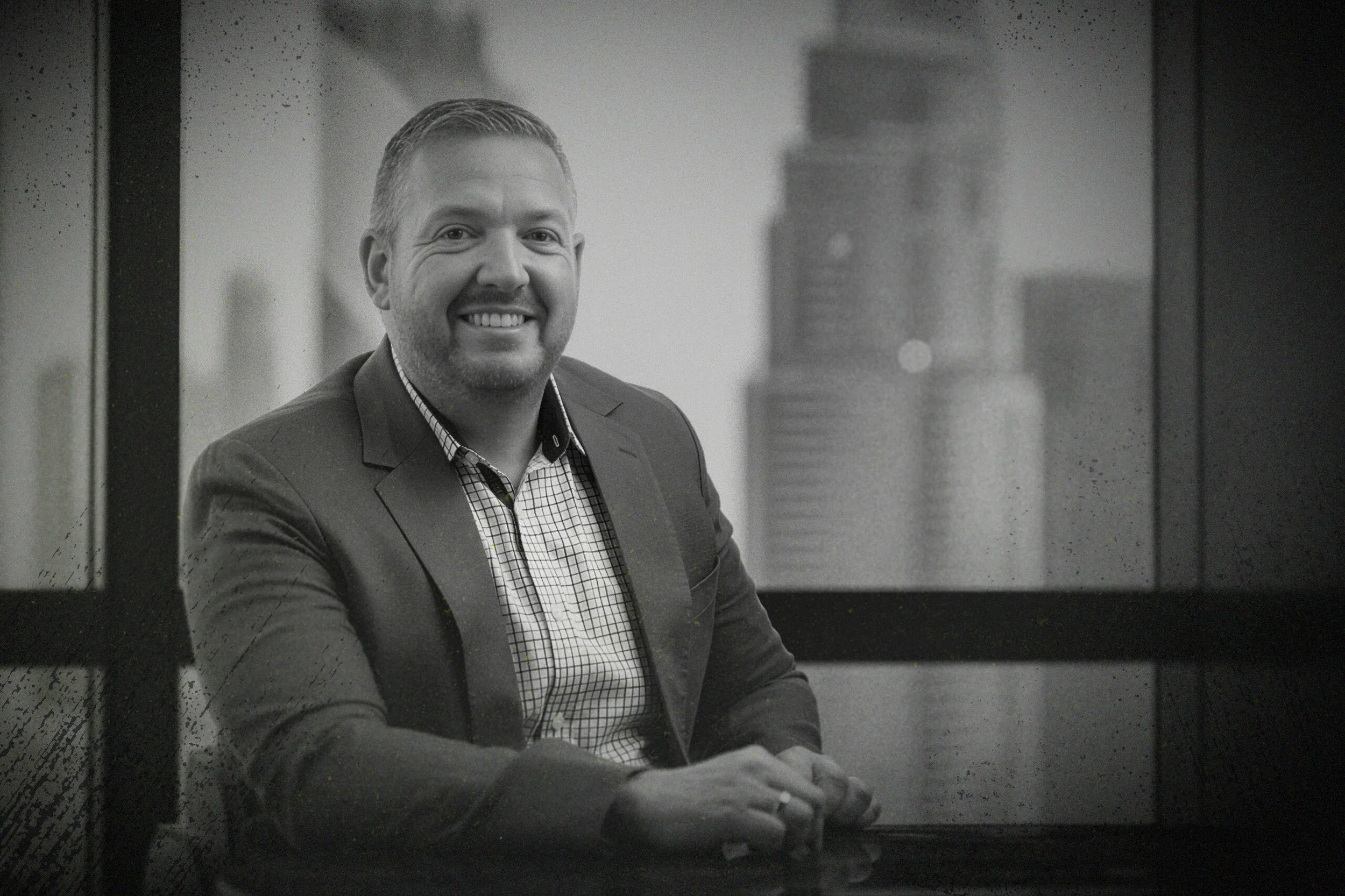 A smiling man in a suit sitting at a table in an office setting with a city skyline visible through the window behind him.