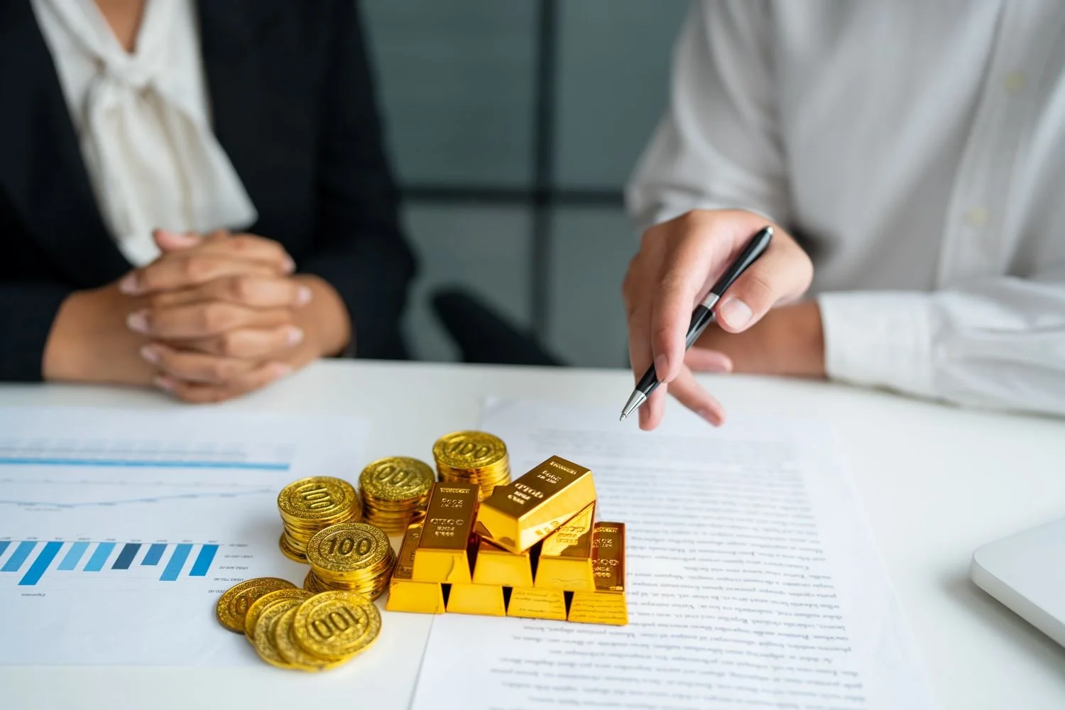 A hand pointing to financial charts topped with stacks of gold bars and coins, symbolizing the high return on investment and business growth that comes from contacting Good as Gold.