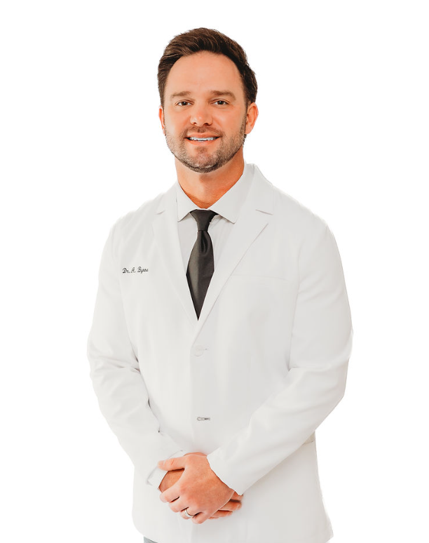 A male doctor wearing a white lab coat, black tie, and smiling against a plain white background.