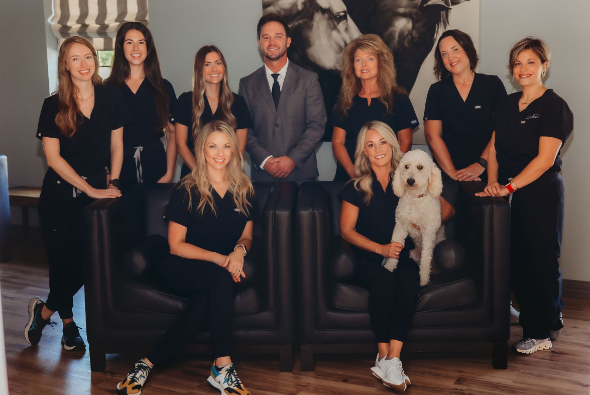 Group photo of nine people, seven women, one man, and a dog, in an indoor setting with framed artwork on the wall.