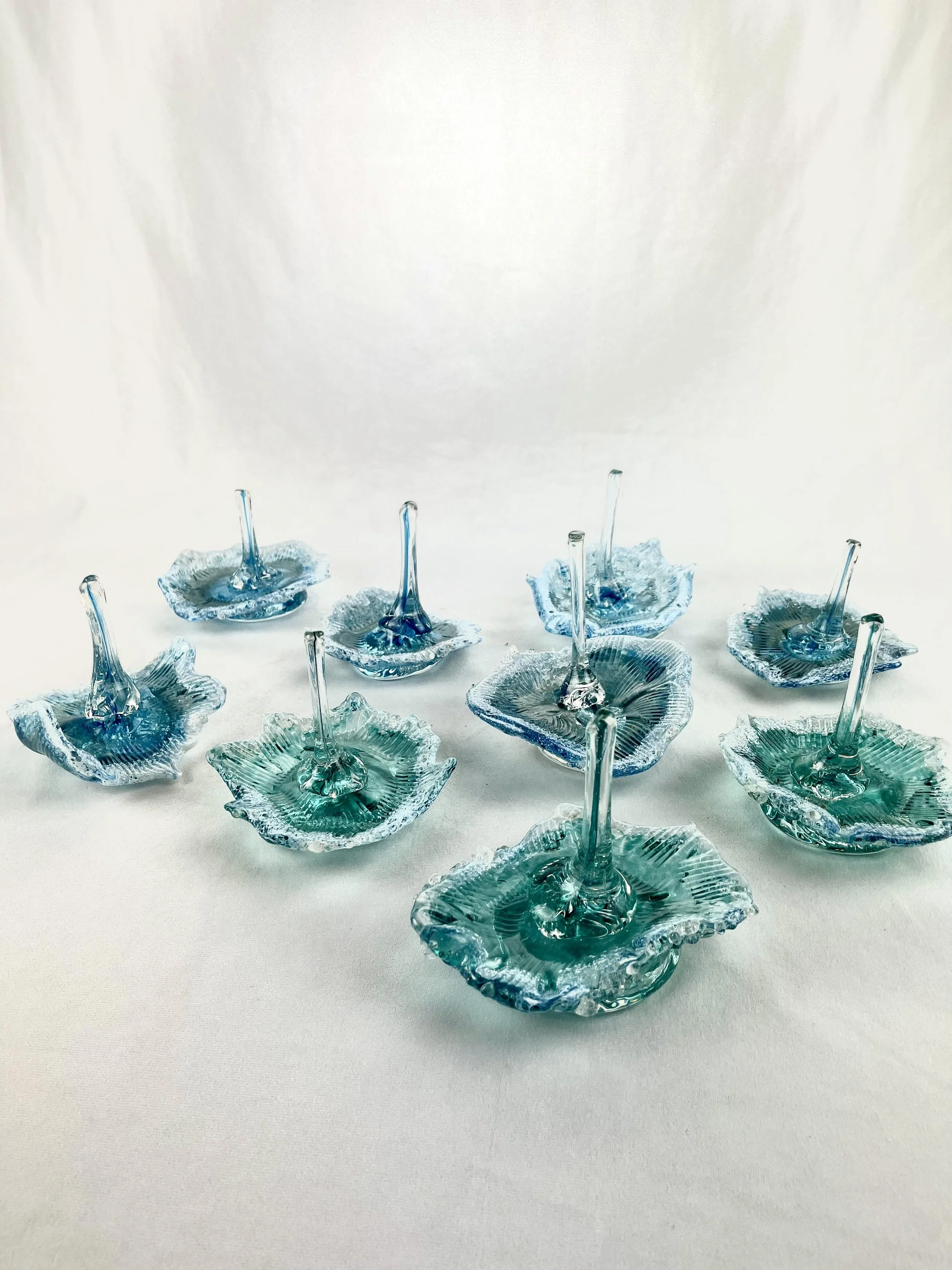 Nine blue and green glass jewelry pieces with elongated stems arranged on a white surface.