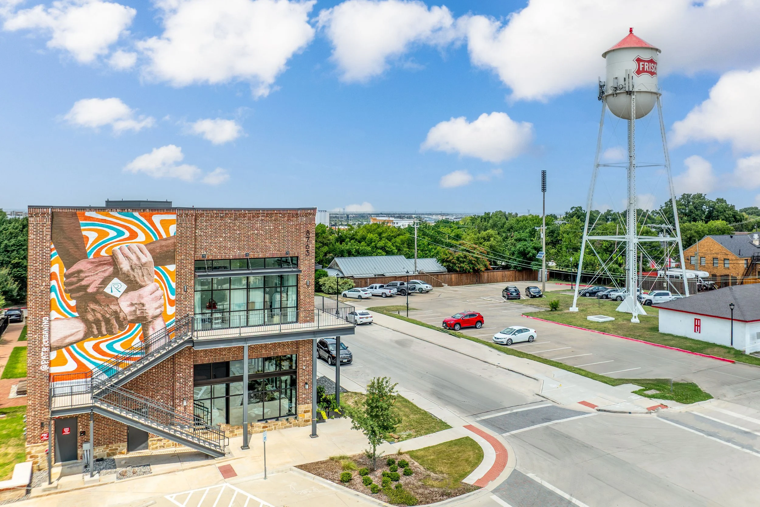 A brick building with a large mural of two hands clasped in solidarity, a parking lot with several cars, a water tower with the Fris logo, and green trees in the background under a partly cloudy blue sky.