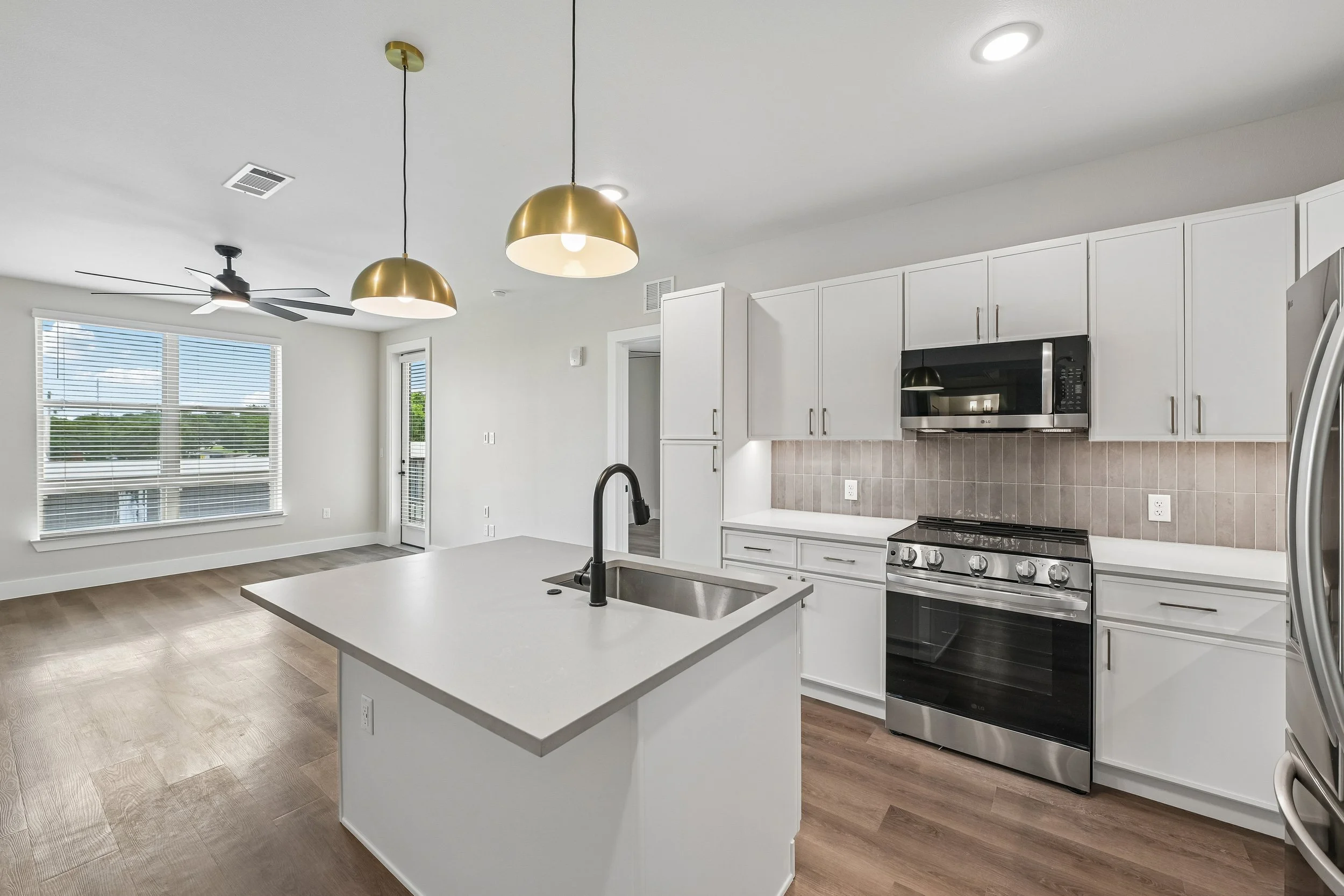 Modern kitchen with white cabinets, stainless steel appliances, a kitchen island with a sink and black faucet, wood flooring, and large window with blinds letting in natural light.