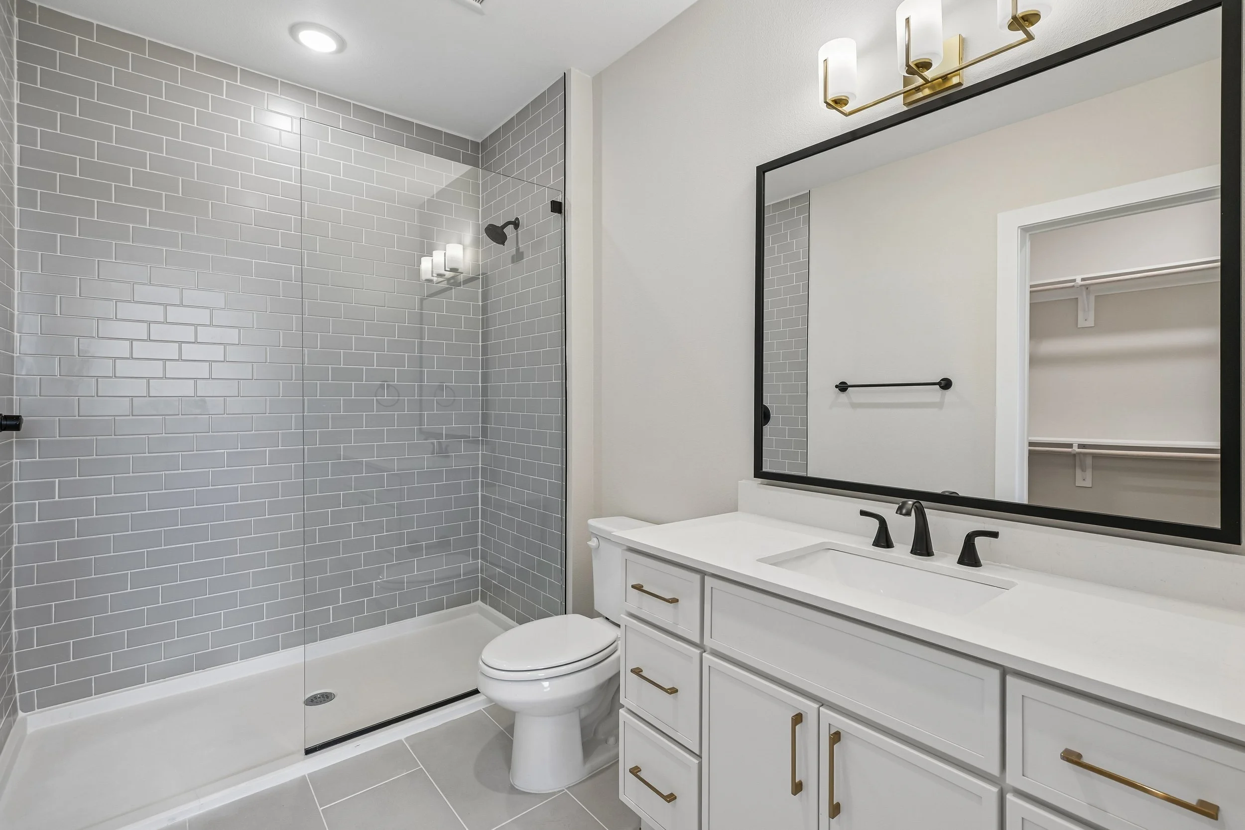 Modern bathroom with a walk-in shower featuring light gray subway tiles, a white vanity with black fixtures, a large mirror, and a small closet or storage area visible through the open doorway.