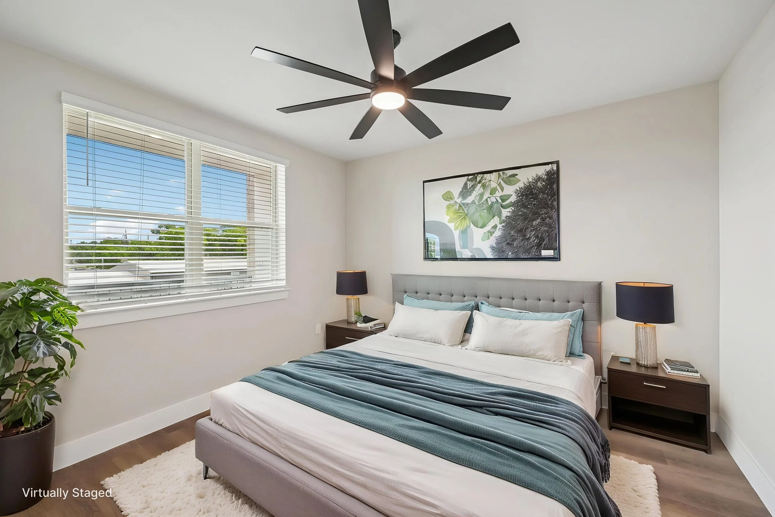 A modern bedroom with a large window, a gray upholstered bed with white and blue bedding, two black bedside lamps, a framed botanical print on the wall, a potted plant in the corner, and a ceiling fan with black blades.
