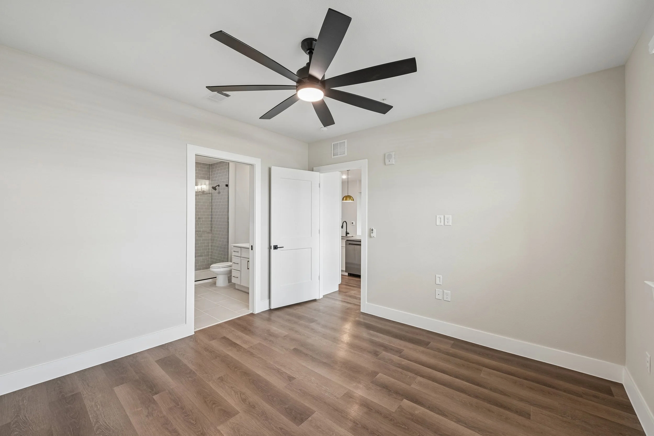 Empty living room with wooden floor, white walls, ceiling fan with black blades, and an open door leading to a bathroom with gray tile shower and a small sink.