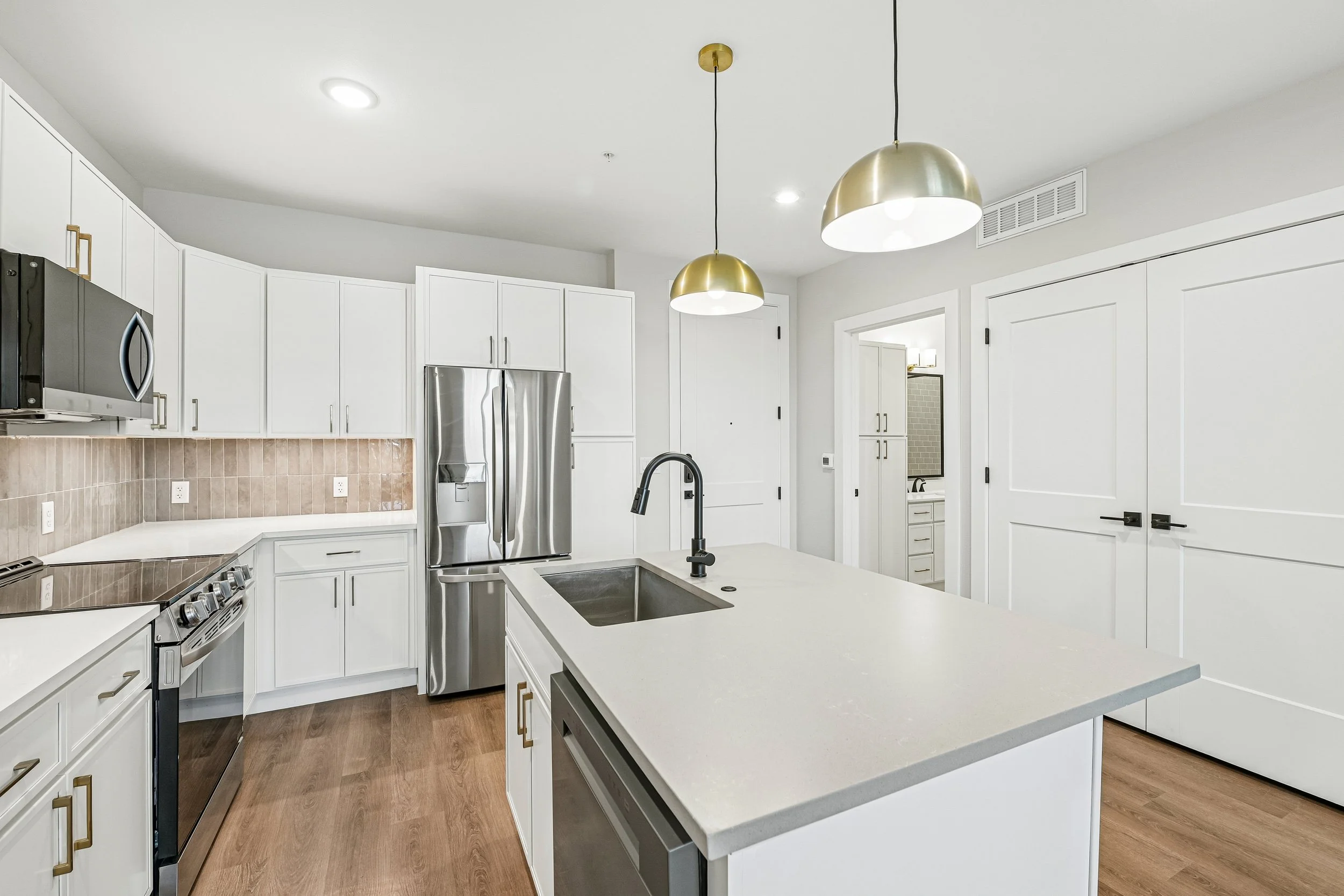 A modern kitchen with white cabinets, a central island with a sink, stainless steel refrigerator, black stove, wood flooring, and gold pendant lighting.