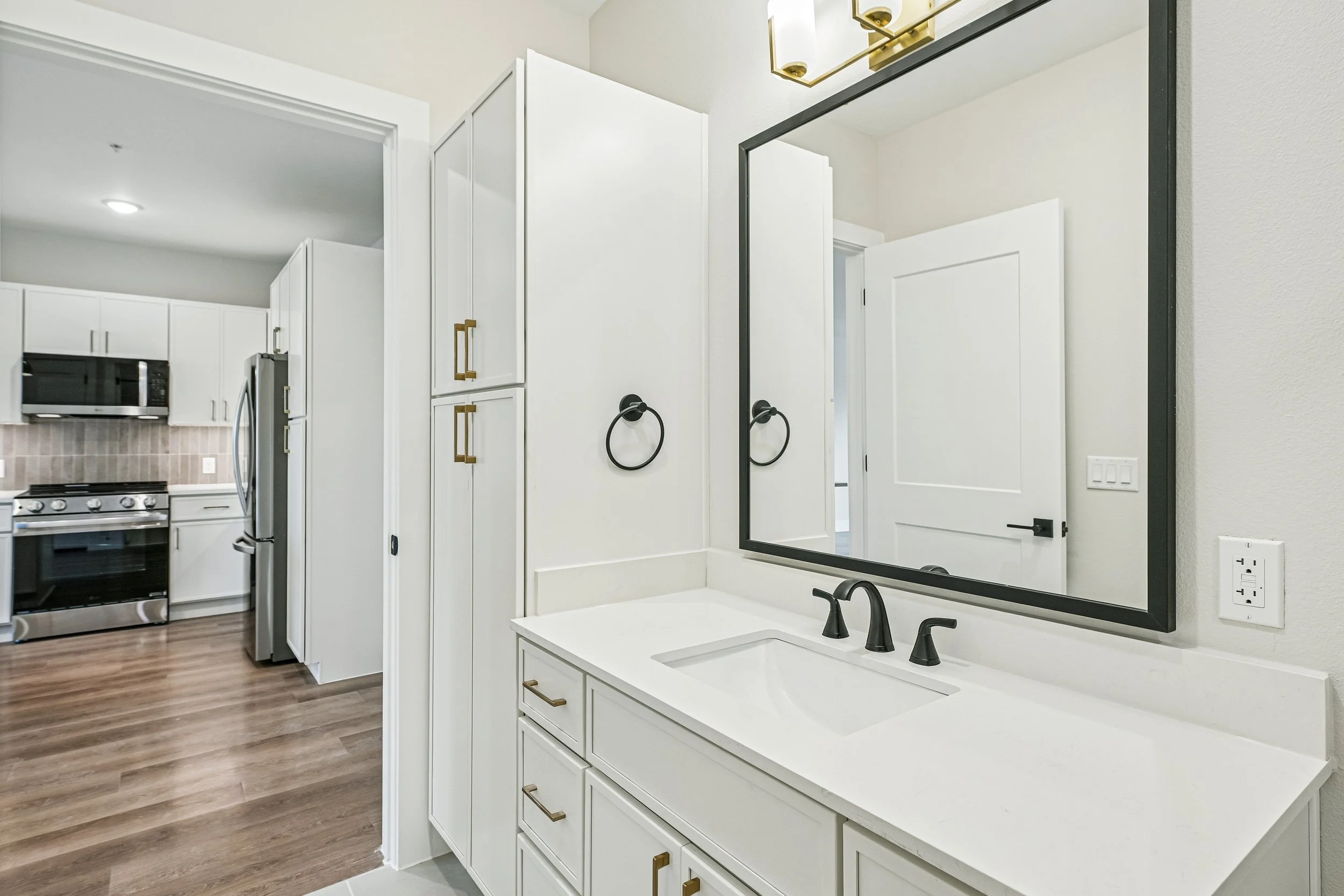 Modern bathroom with white vanity, black fixtures, large wall mirror, gold and black light fixture, and a view into a kitchen area with white cabinets and stainless steel refrigerator.
