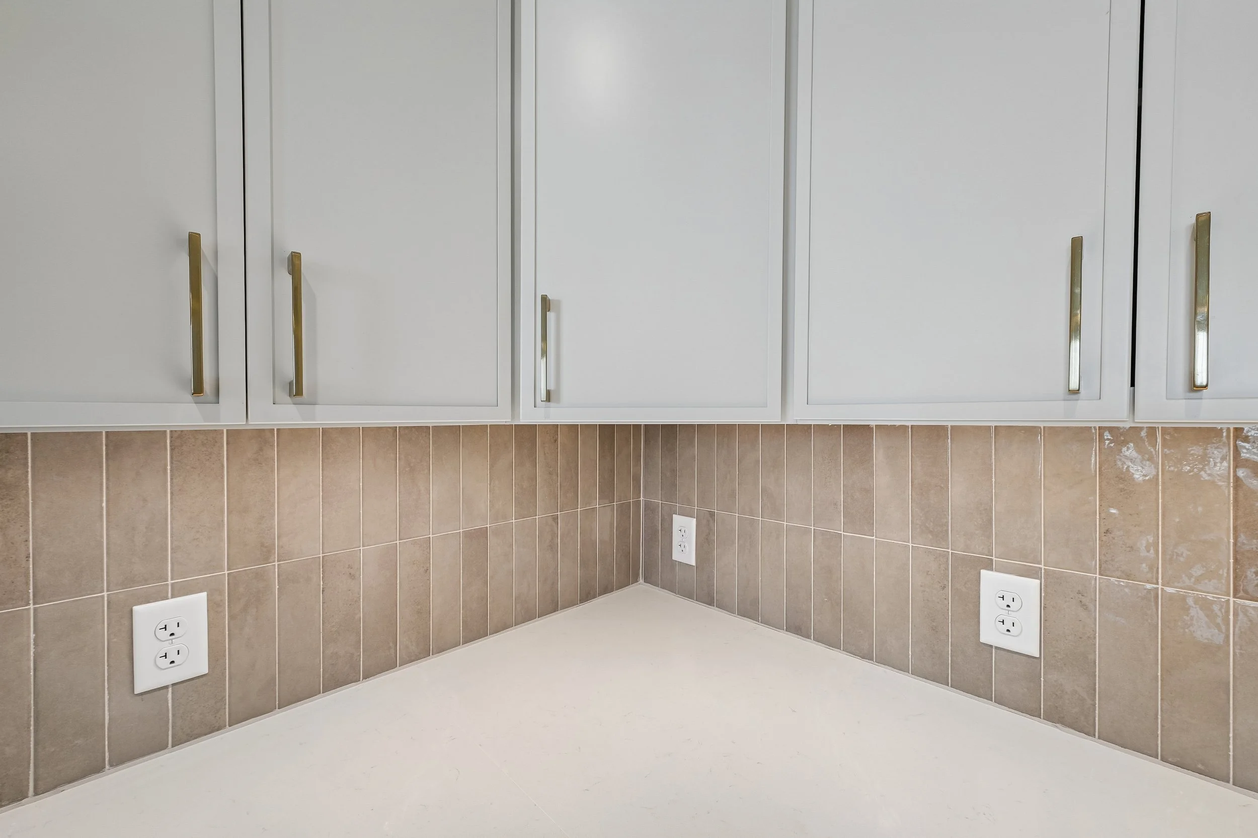 White kitchen cabinets with gold handles above a beige tiled backsplash and four white electrical outlets.