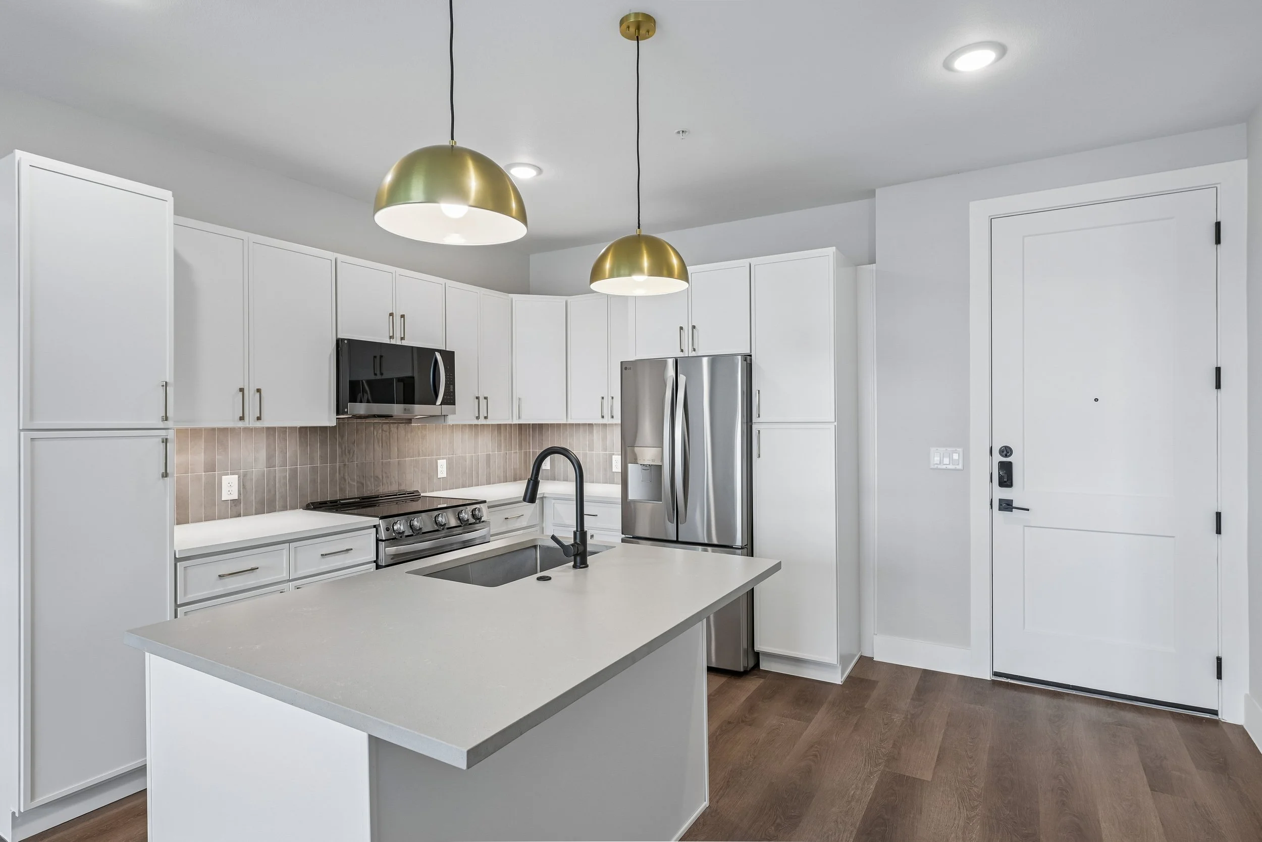 Modern kitchen with white cabinets, stainless steel refrigerator, black microwave, black faucet, beige tiled backsplash, white island with a countertop, and wooden flooring. Two gold pendant lights hang from the ceiling.