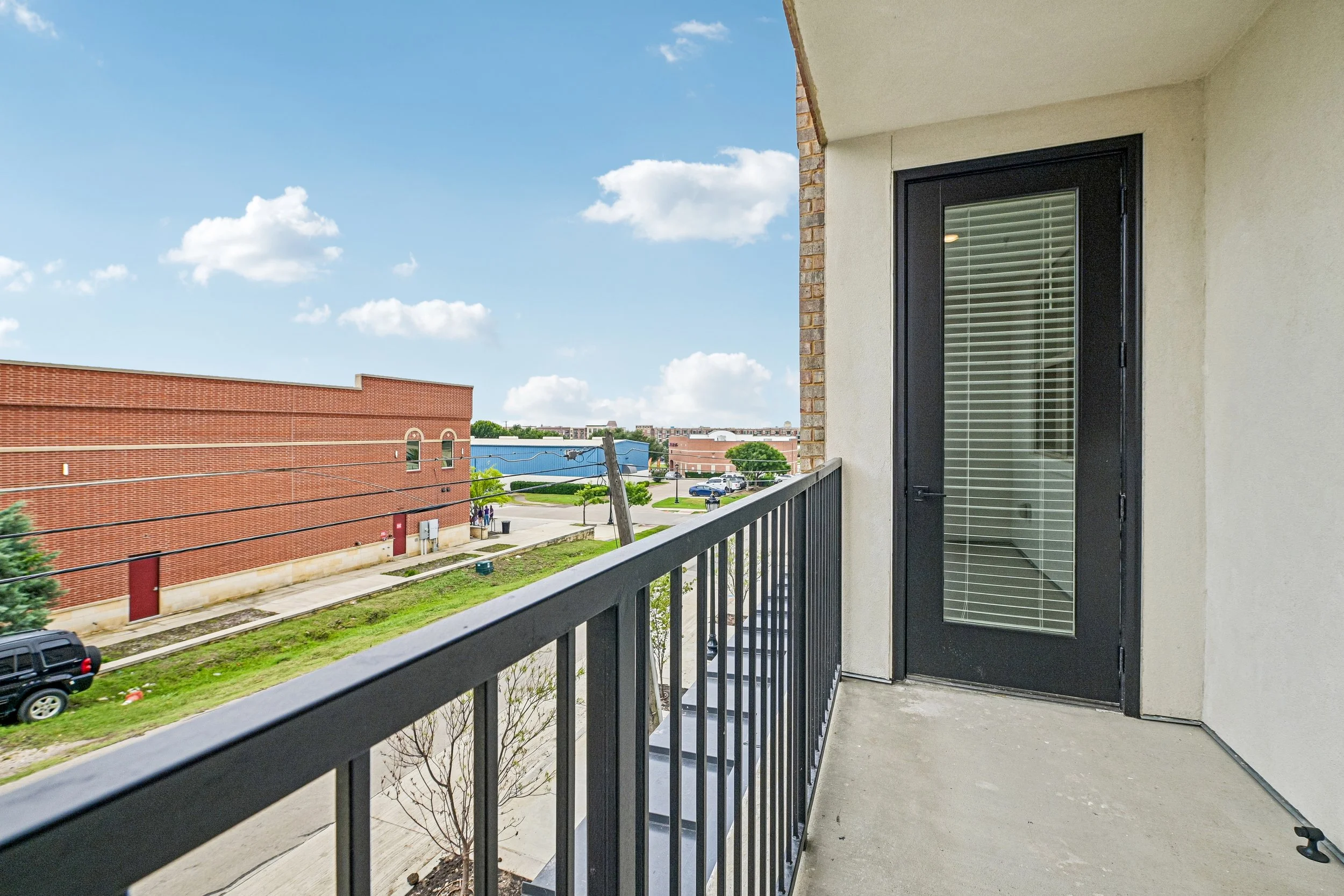 View from apartment balcony with black railing and glass door, overlooking a parking lot, red brick building, and blue sky with clouds.
