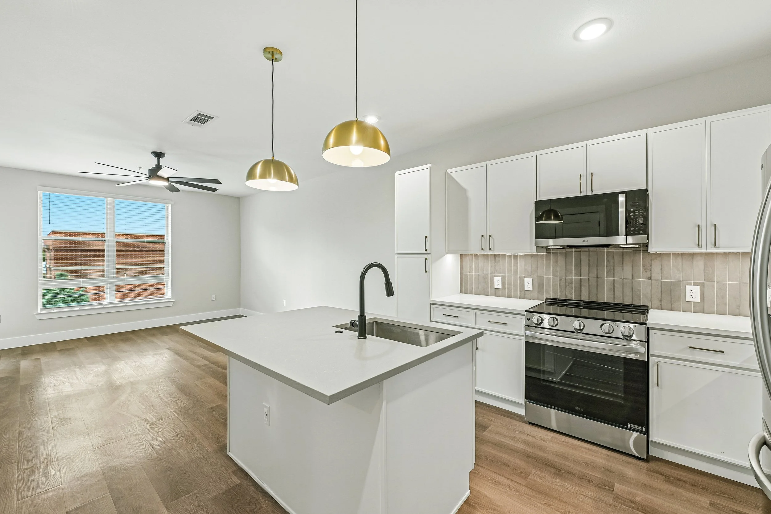 Modern kitchen with white cabinets, stainless steel oven, microwave, black faucet, and gold pendant lights, with a large window and beige tile backsplash.