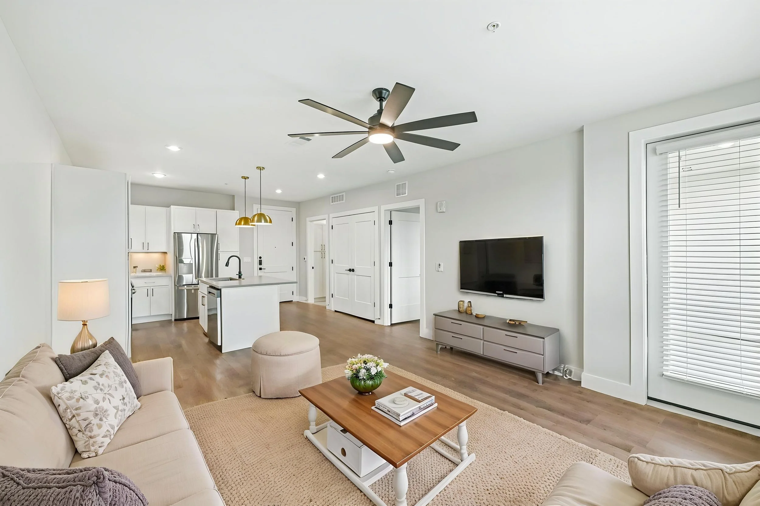 Living room with beige sofa, coffee table, wall-mounted TV, white cabinets, kitchen area with island, pendant lights, ceiling fan, and large window with blinds.