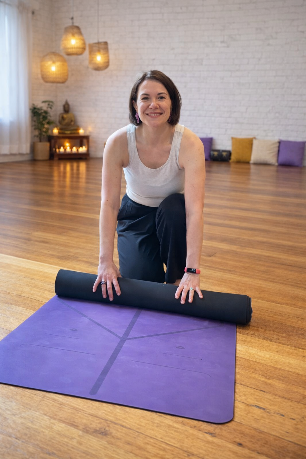 A woman preparing for yoga on a purple mat in a bright, cozy studio with candles and a Buddha statue in the background.
