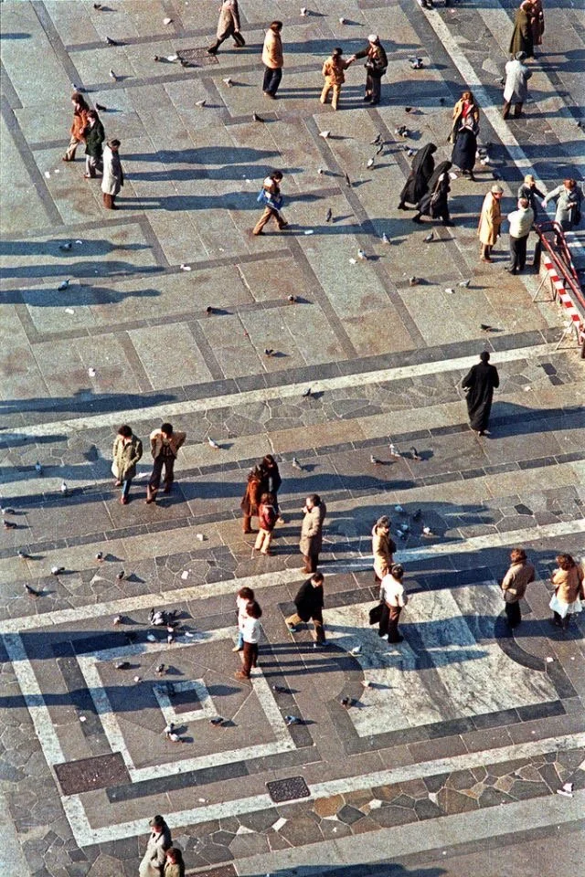 People walking across a large tiled public square with pigeons on the ground.