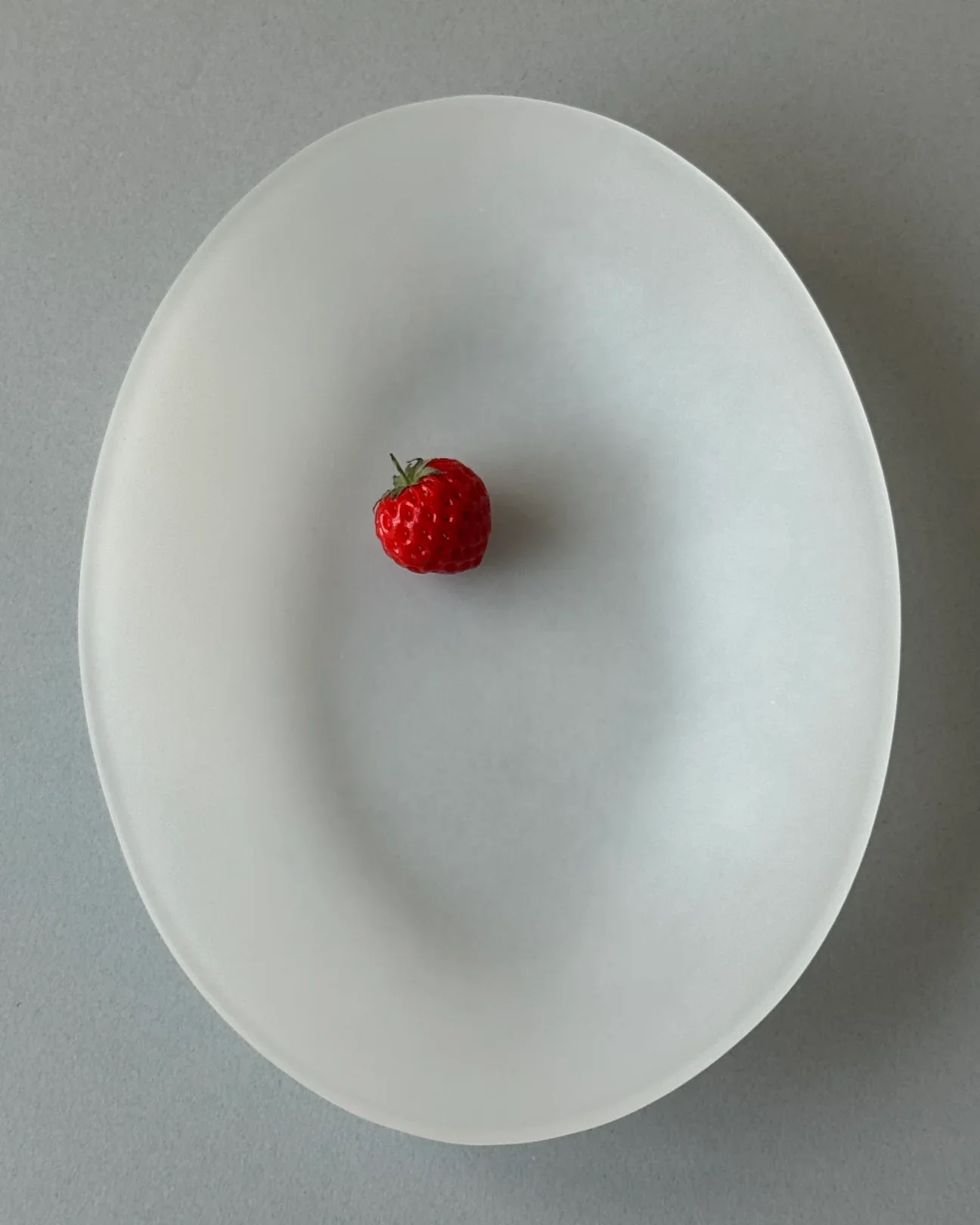 A single red strawberry with a green stem on a white oval-shaped plate against a light gray background.