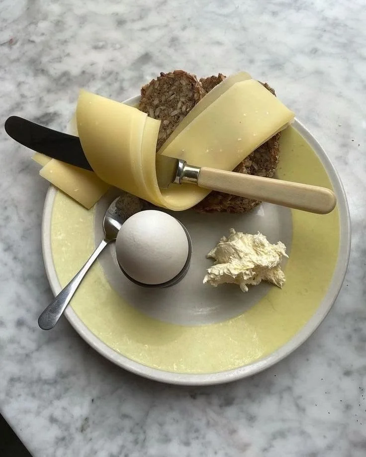 A plate of cheese, bread, butter, egg, and a knife with a black handle, on a marble surface.