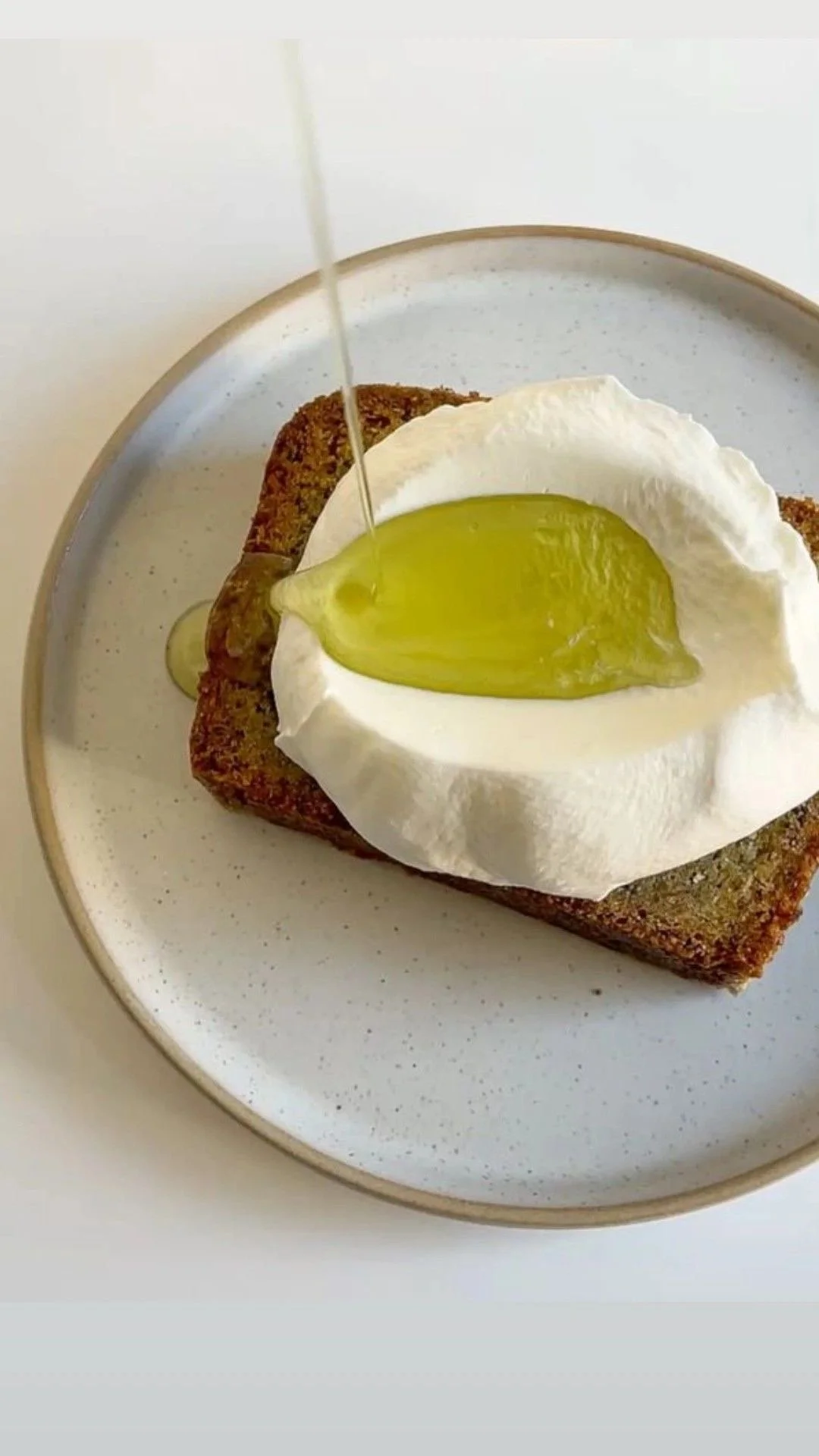 Slice of bread topped with a dollop of white cream cheese, a slice of pickled cucumber, and a piece of fried bread, with a stream of honey pouring over it, on a white plate.